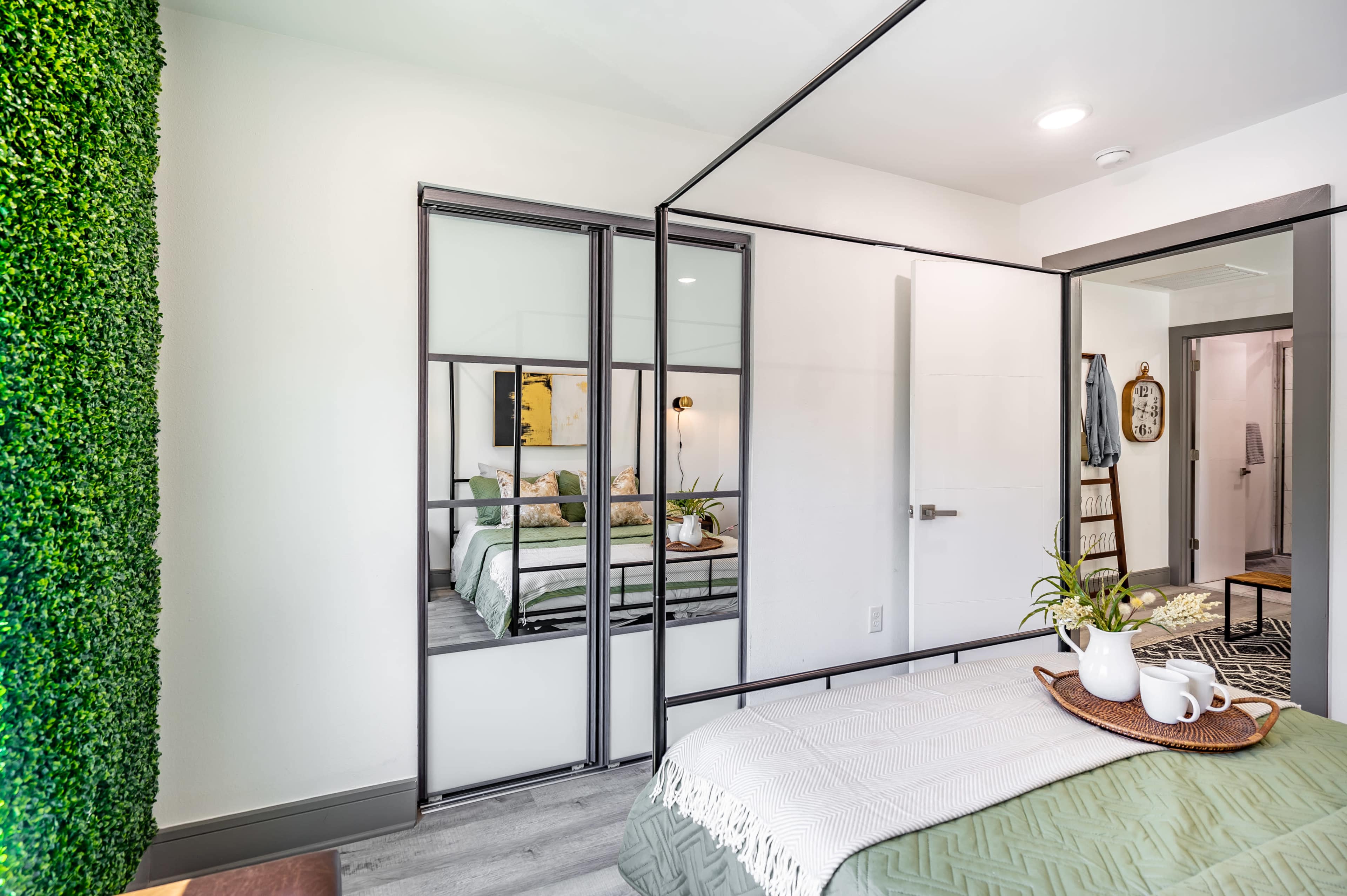 A modern bedroom with a glass-paneled wardrobe, a bed with decorative pillows, and a green moss wall element in one corner.