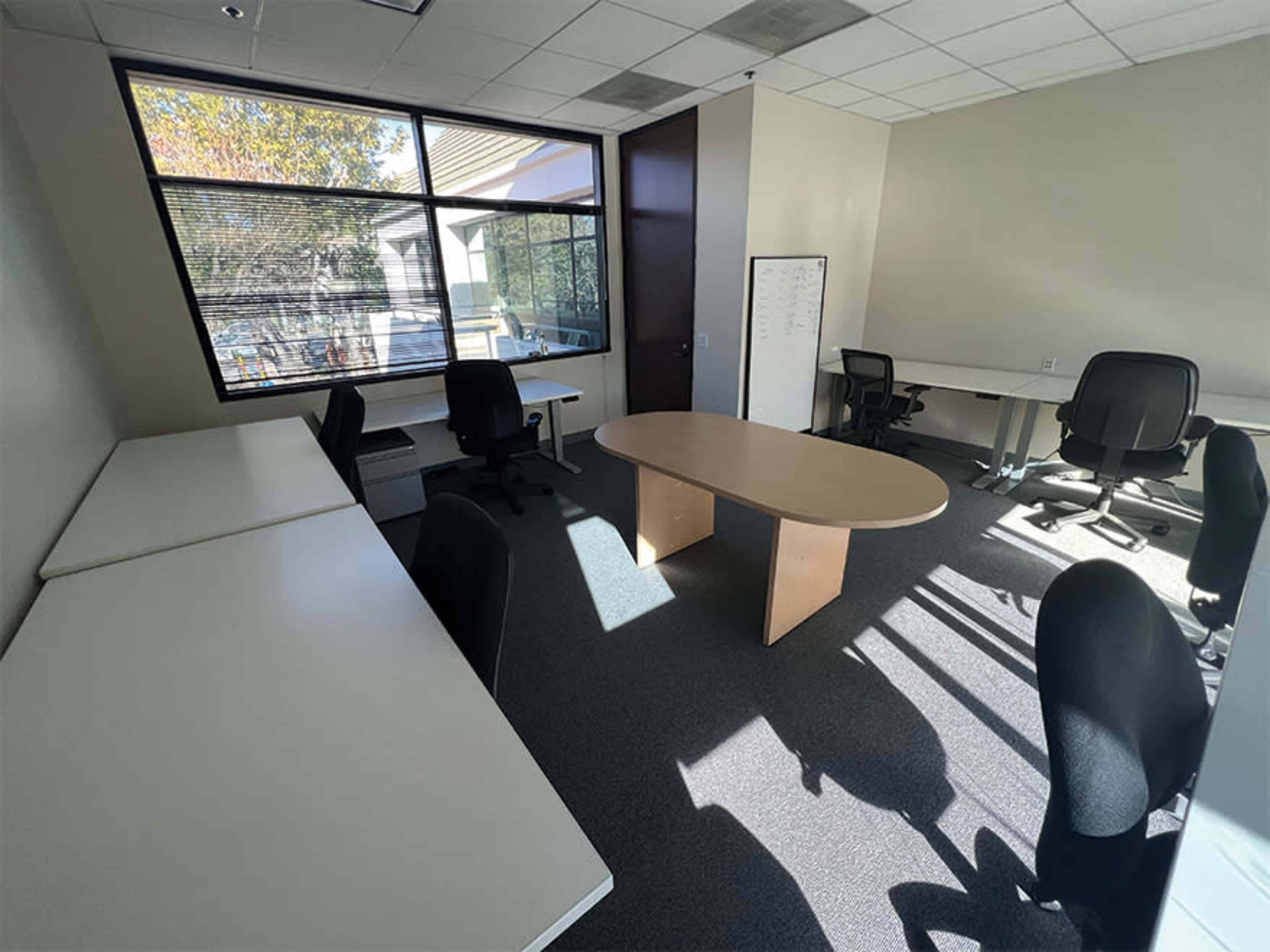 An office space with four black chairs, a large oval table, and two desks positioned against the walls, illuminated by natural light from a window.
