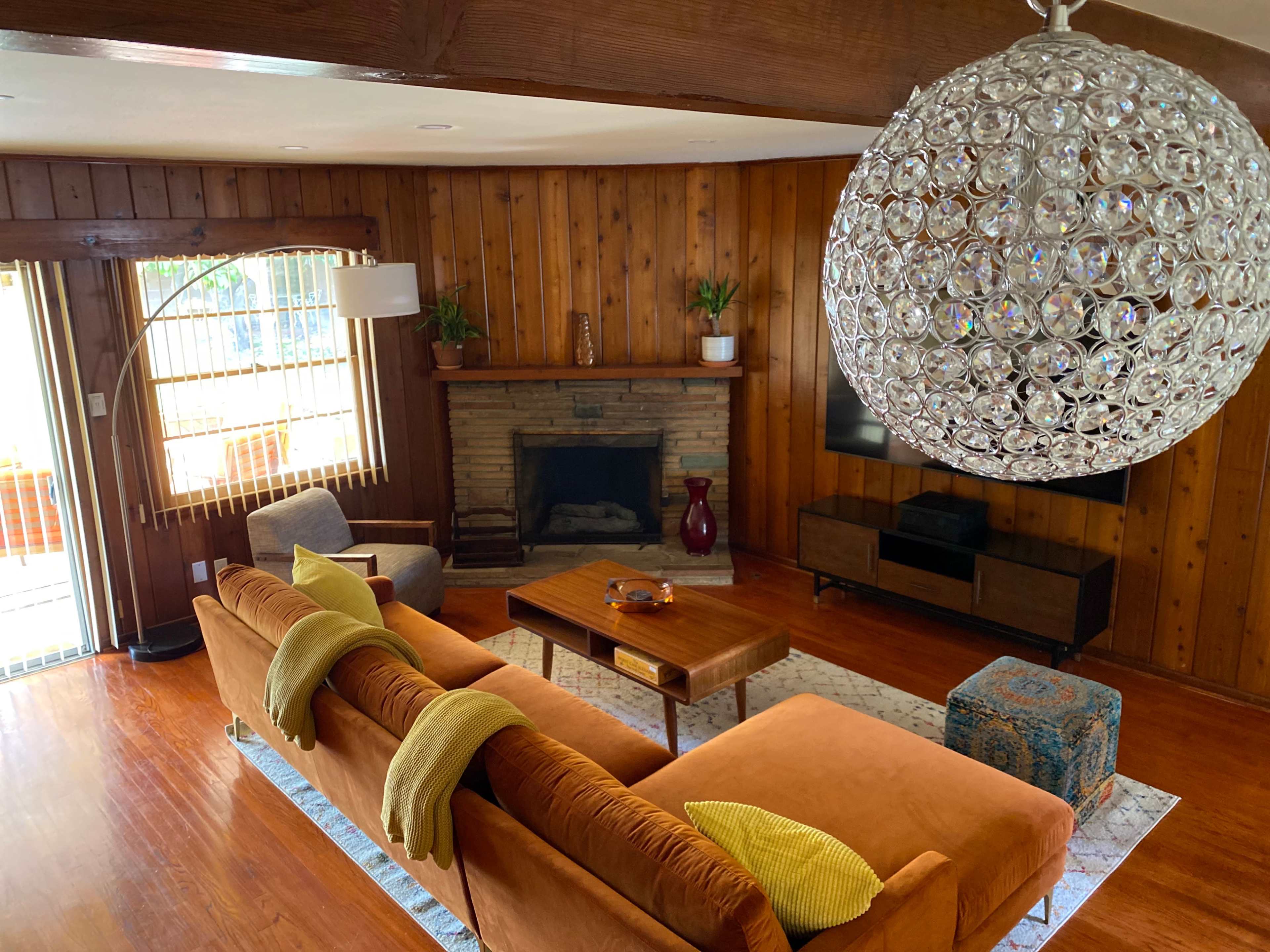 A spacious living room with wood paneling, a stone fireplace, a large round chandelier, and a sofa adorned with yellow throw blankets.