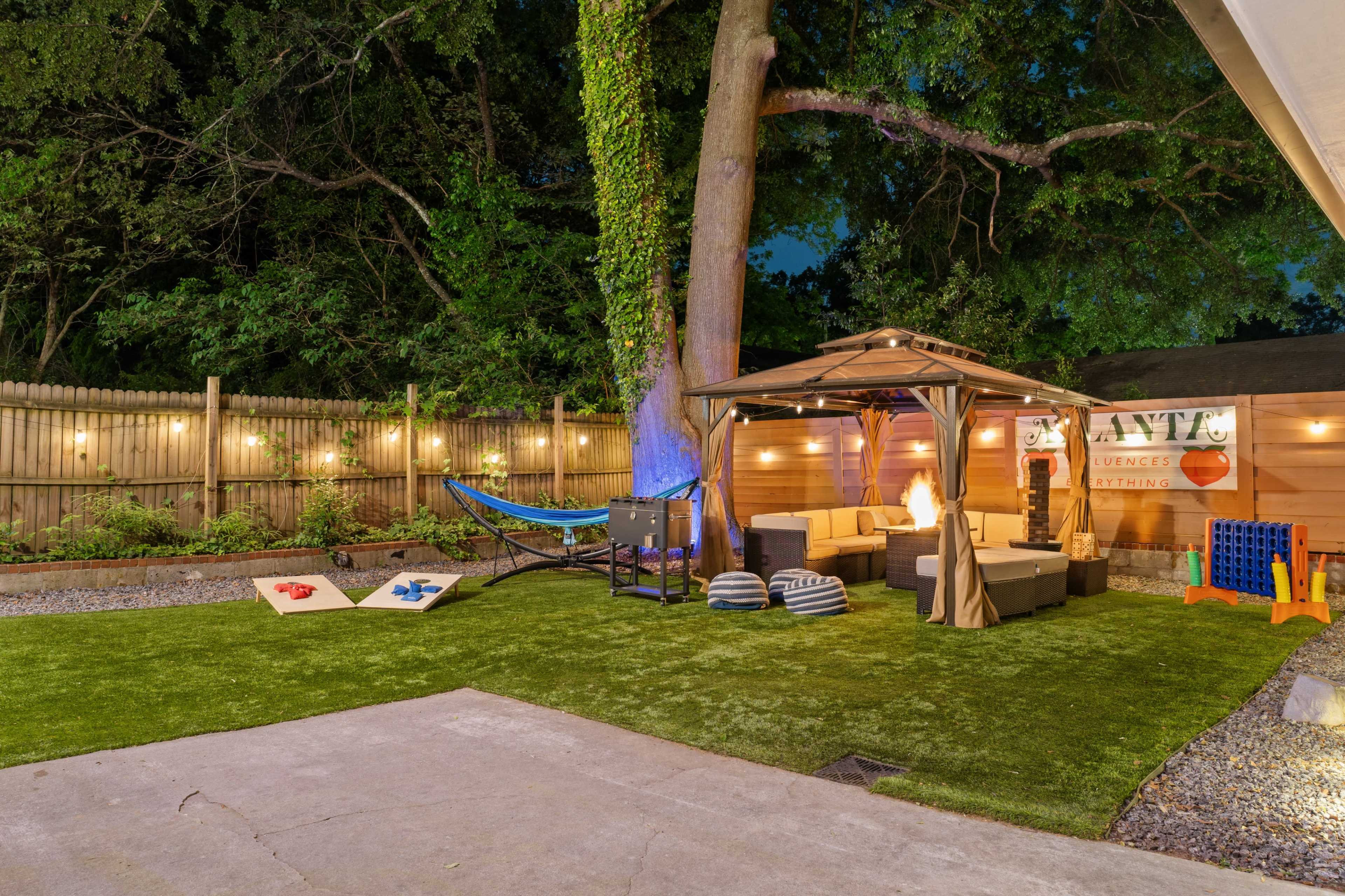 The image shows a backyard scene featuring a seating area under a canopy, a hammock, outdoor games, and decorative lighting among trees and a wooden fence.