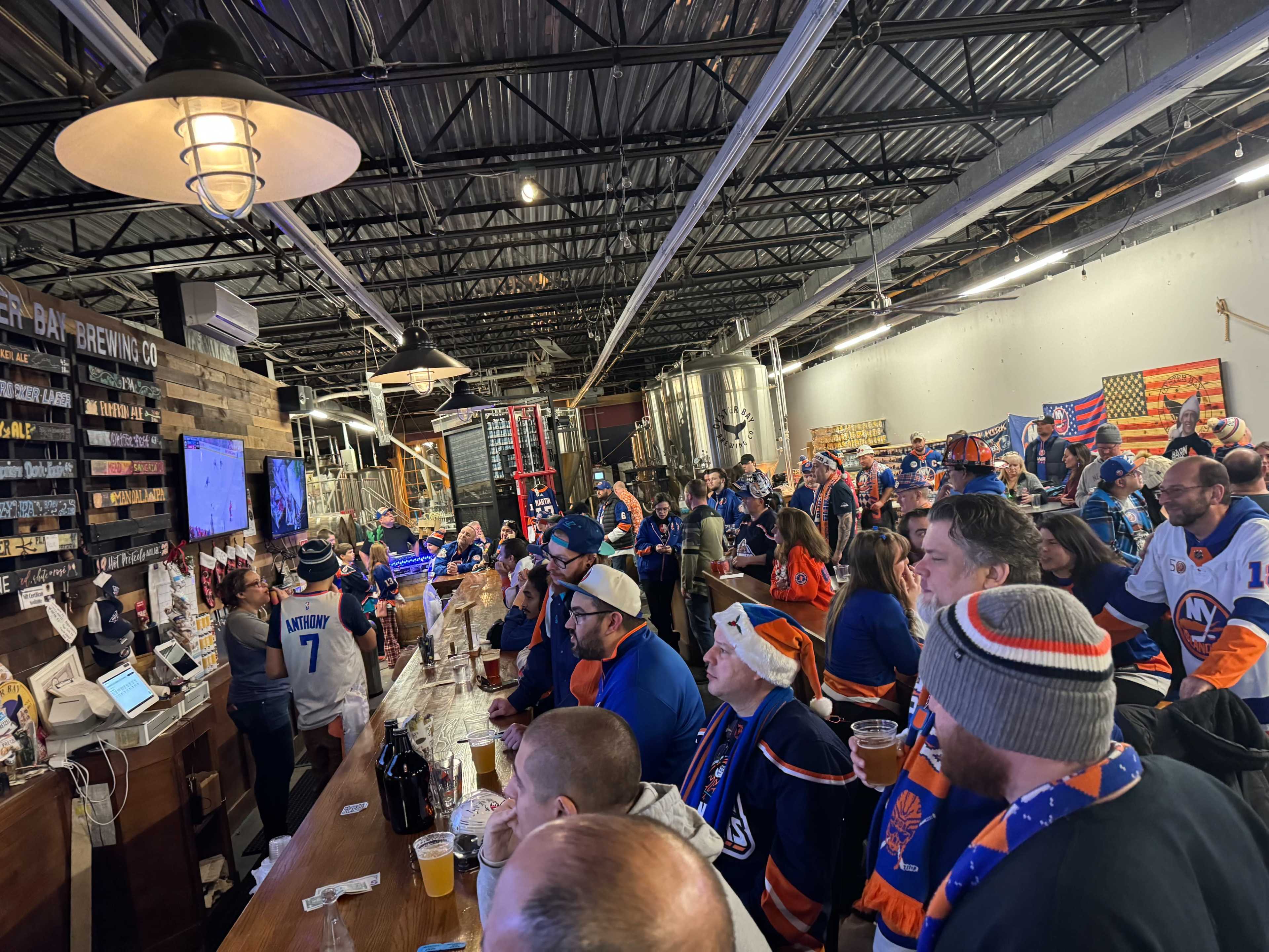 A large crowd of sports fans wearing team jerseys and scarves gathers at a bar to watch a game on a big screen.