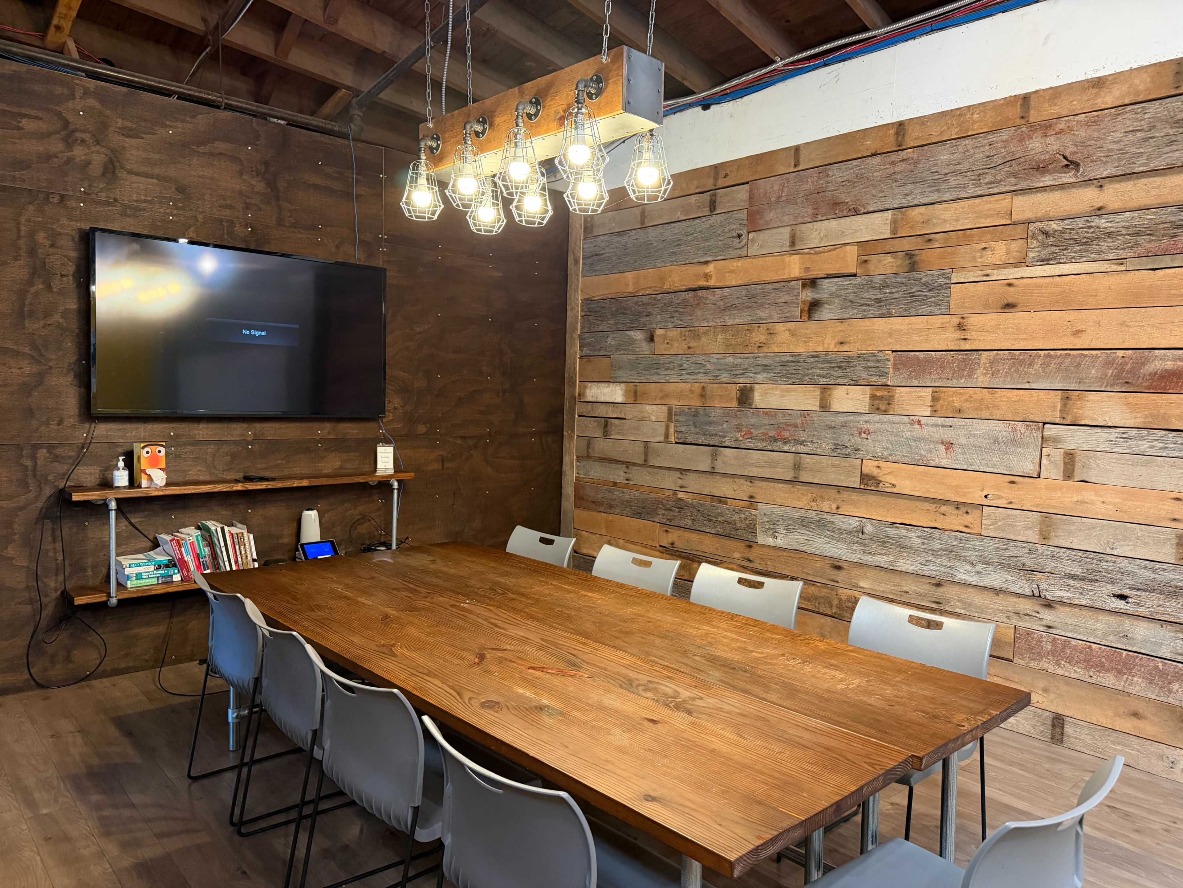 The image shows a conference room with a wooden table, several gray chairs, a large television mounted on a wall, and a rustic wooden panel feature.