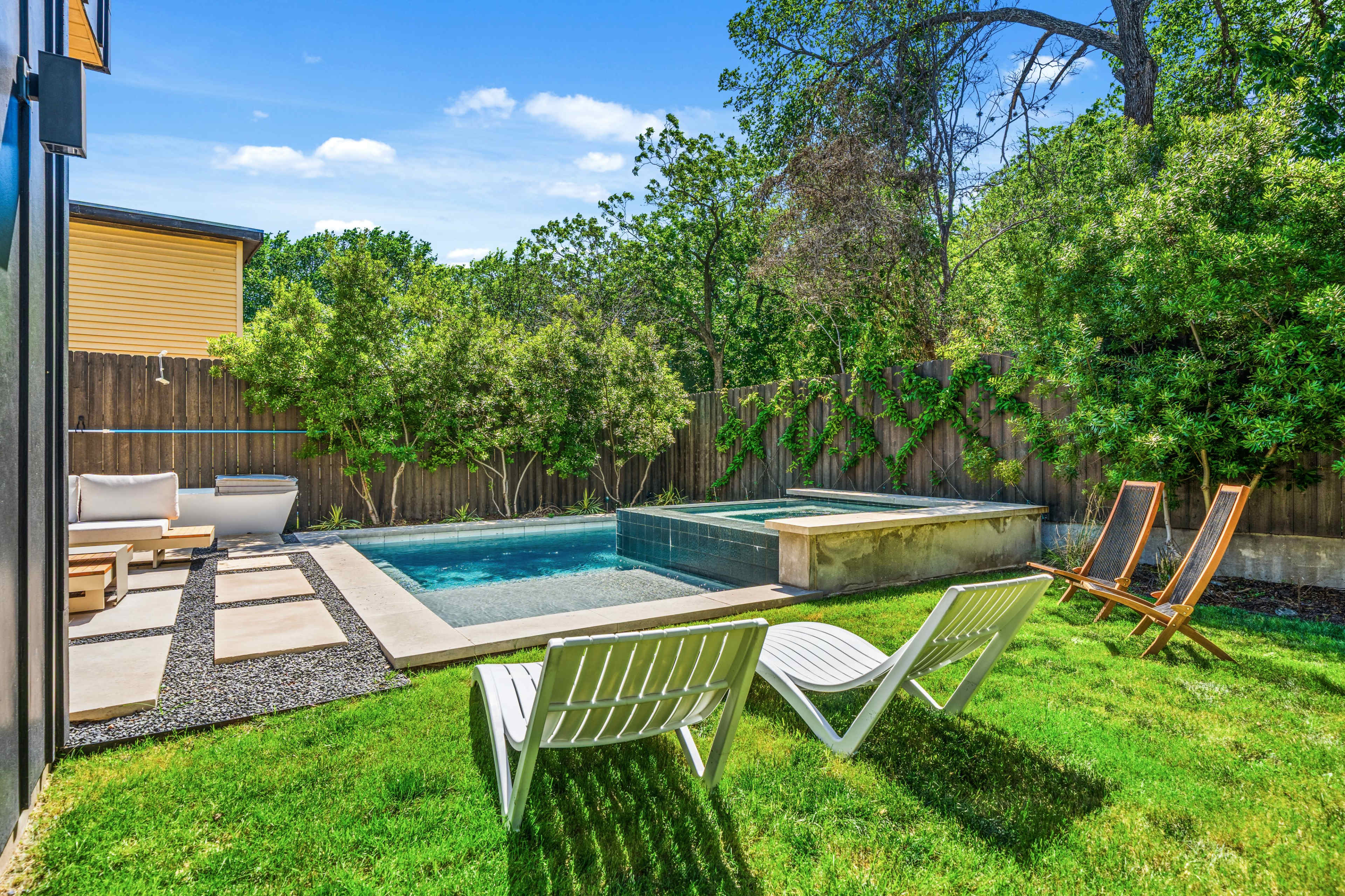 The image shows a backyard with a swimming pool, a hot tub, two lounge chairs, and a grassy area surrounded by trees and a wooden fence.