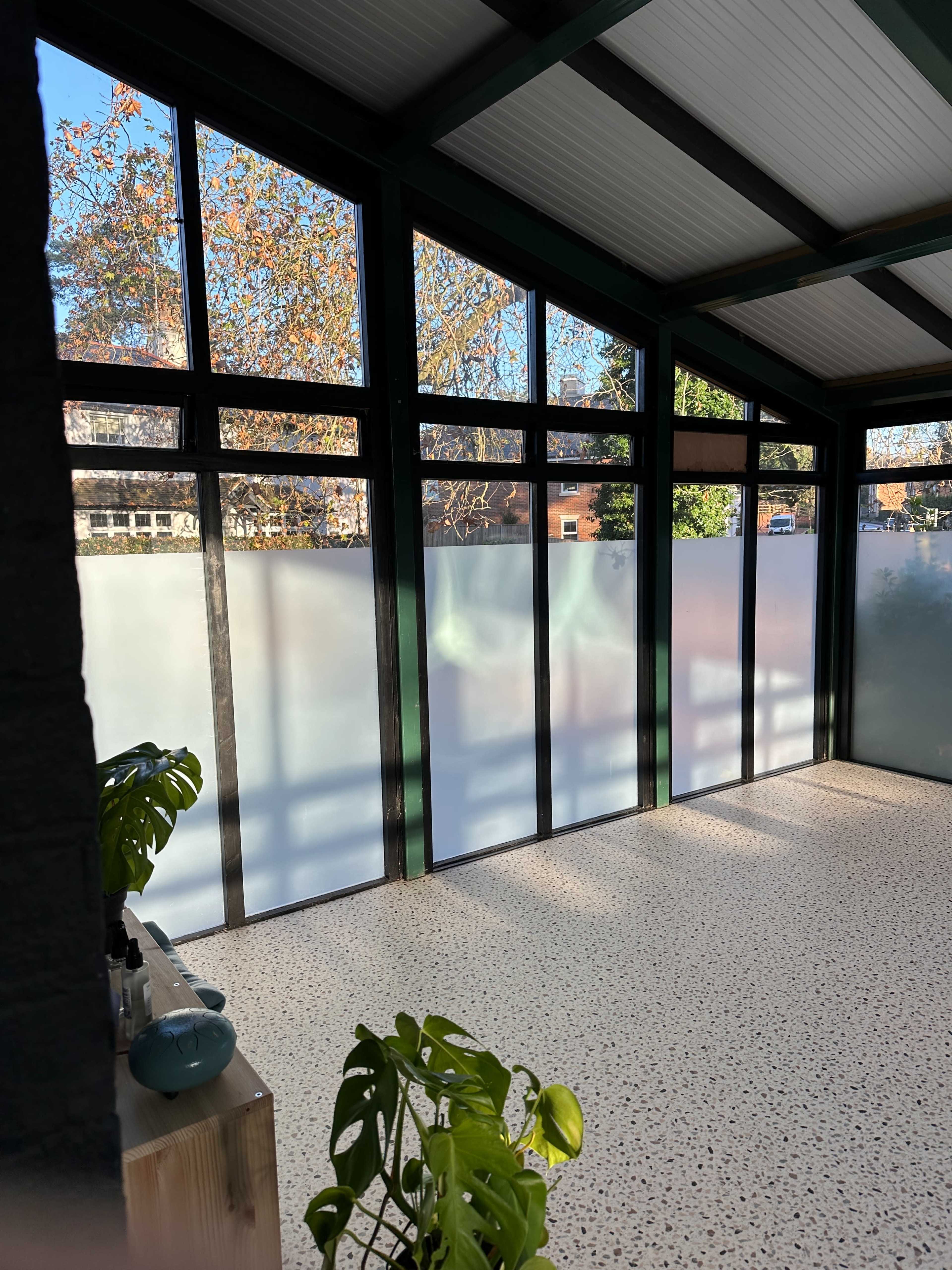 The image shows a sunlit indoor space featuring large glass windows, a light-colored floor, and plants placed near the wall.