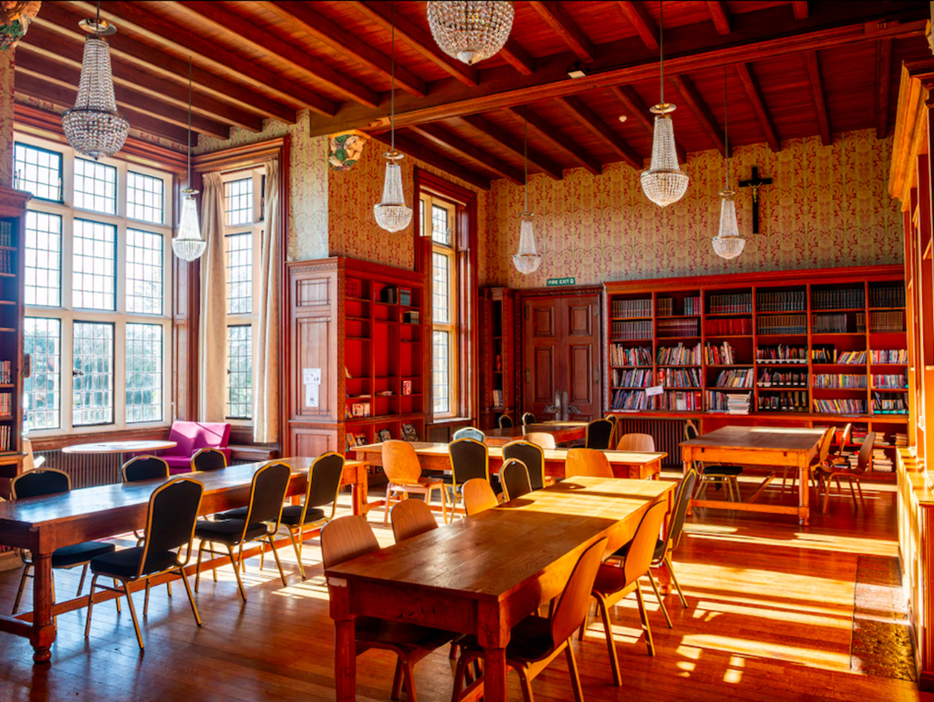 A bright, spacious library with wooden tables and chairs arranged around a room filled with bookshelves and large windows allowing natural light to enter.