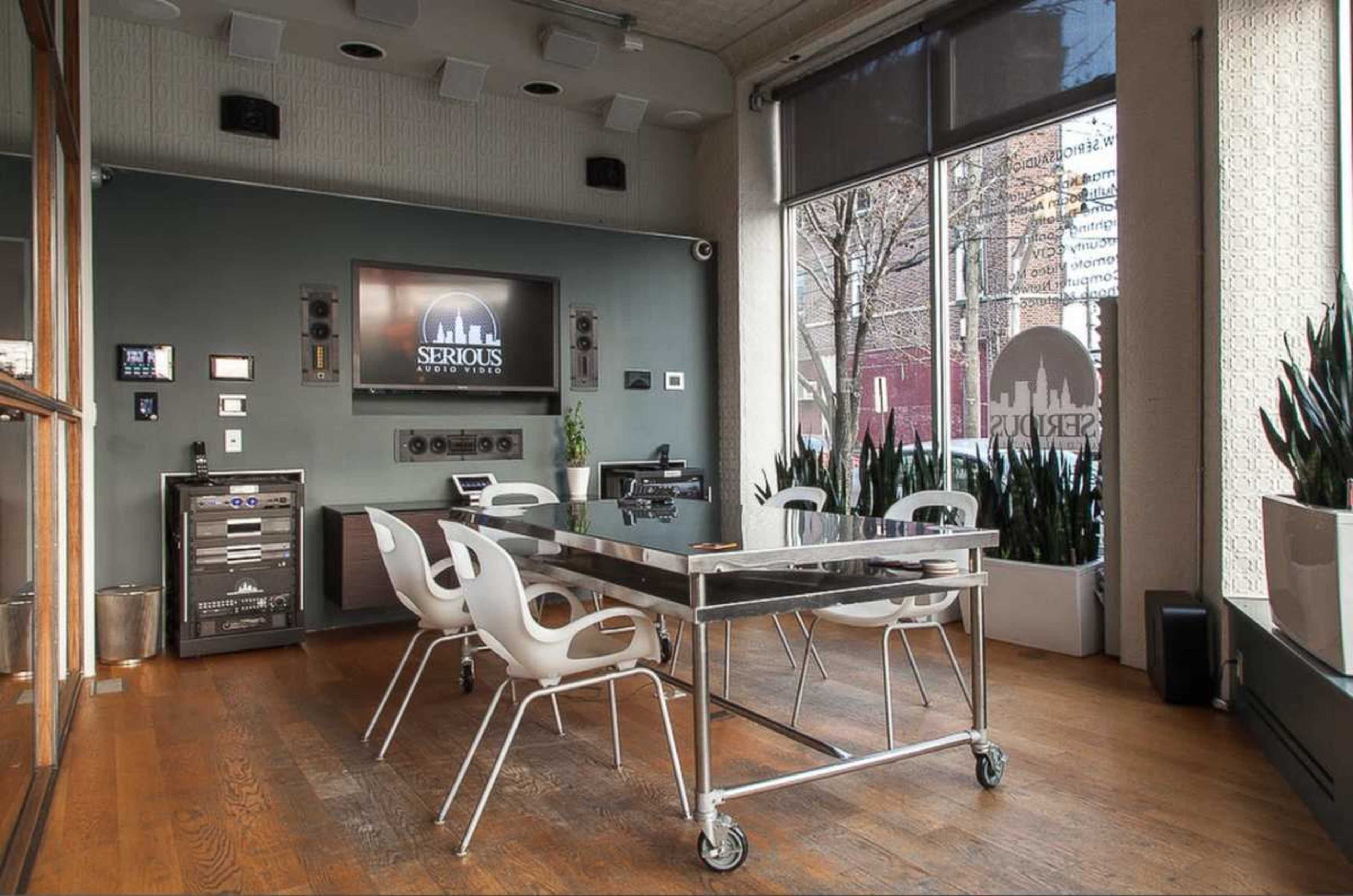A modern conference room features a sleek glass table surrounded by white chairs, with a large screen and various audio-visual equipment on the walls.