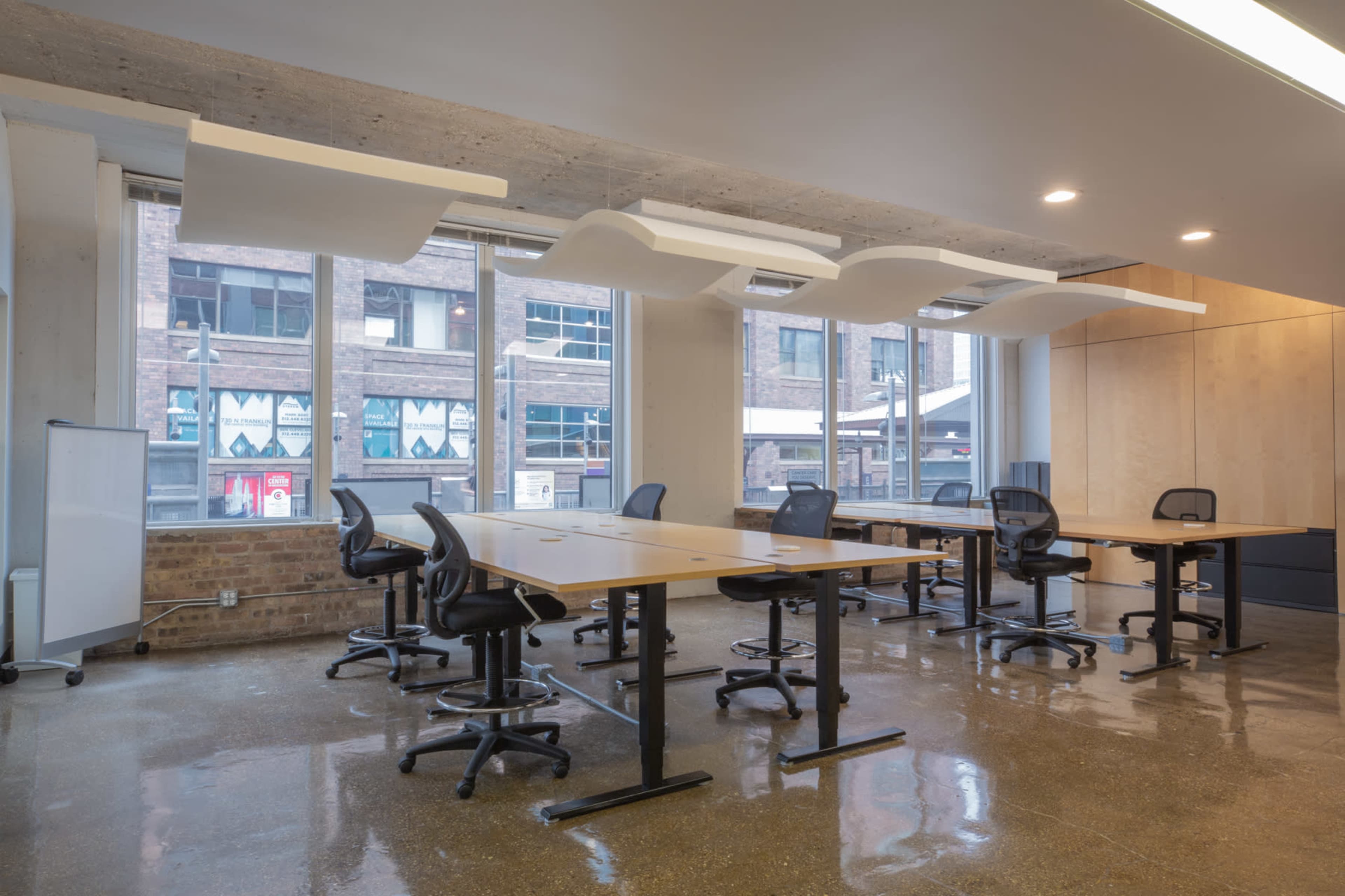 The image depicts a modern office space with multiple desks and ergonomic chairs arranged in an open layout, featuring large windows that allow natural light to enter.