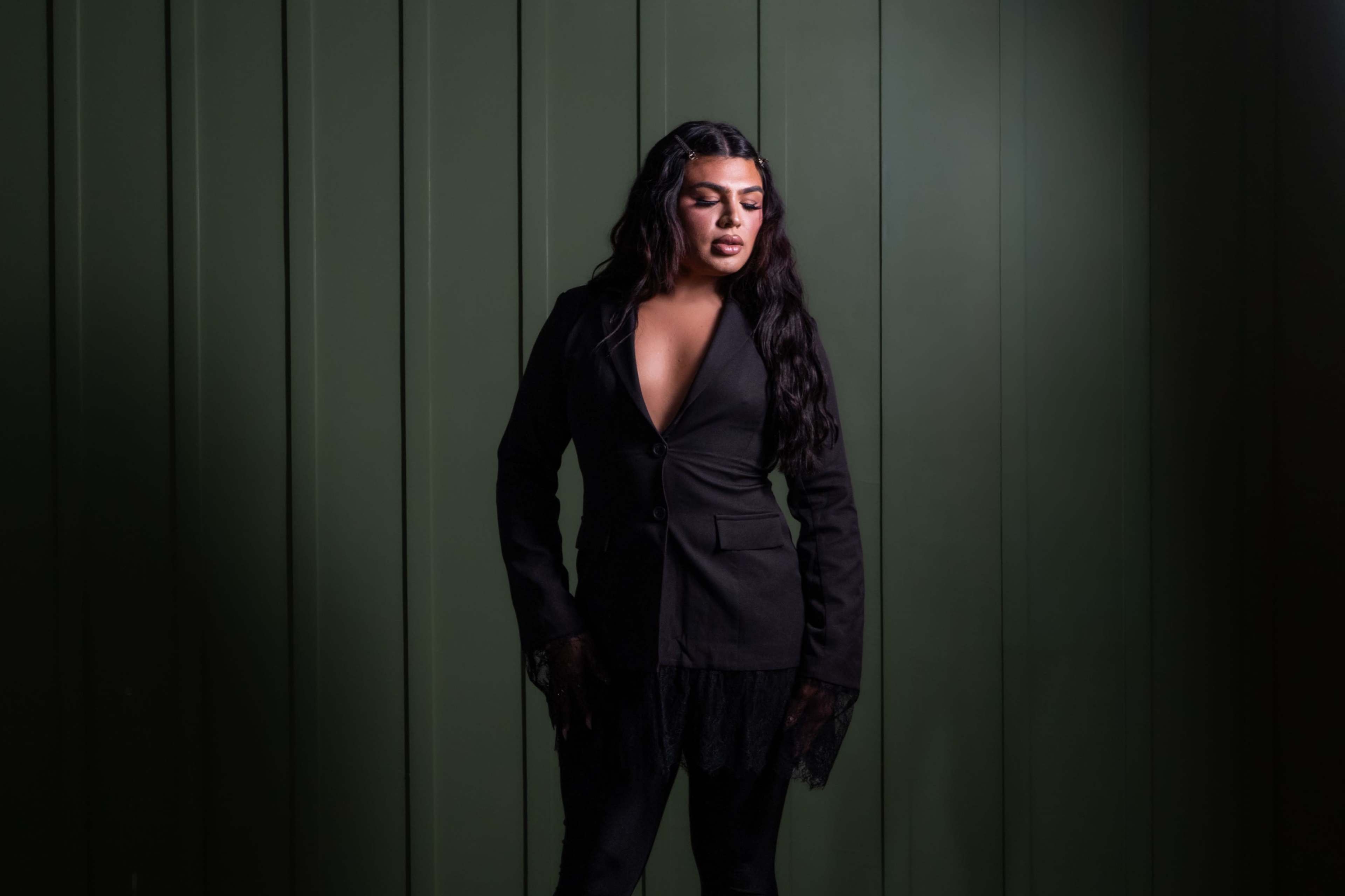 A person stands against a green wall, wearing a black suit jacket with lace details and styled long hair.