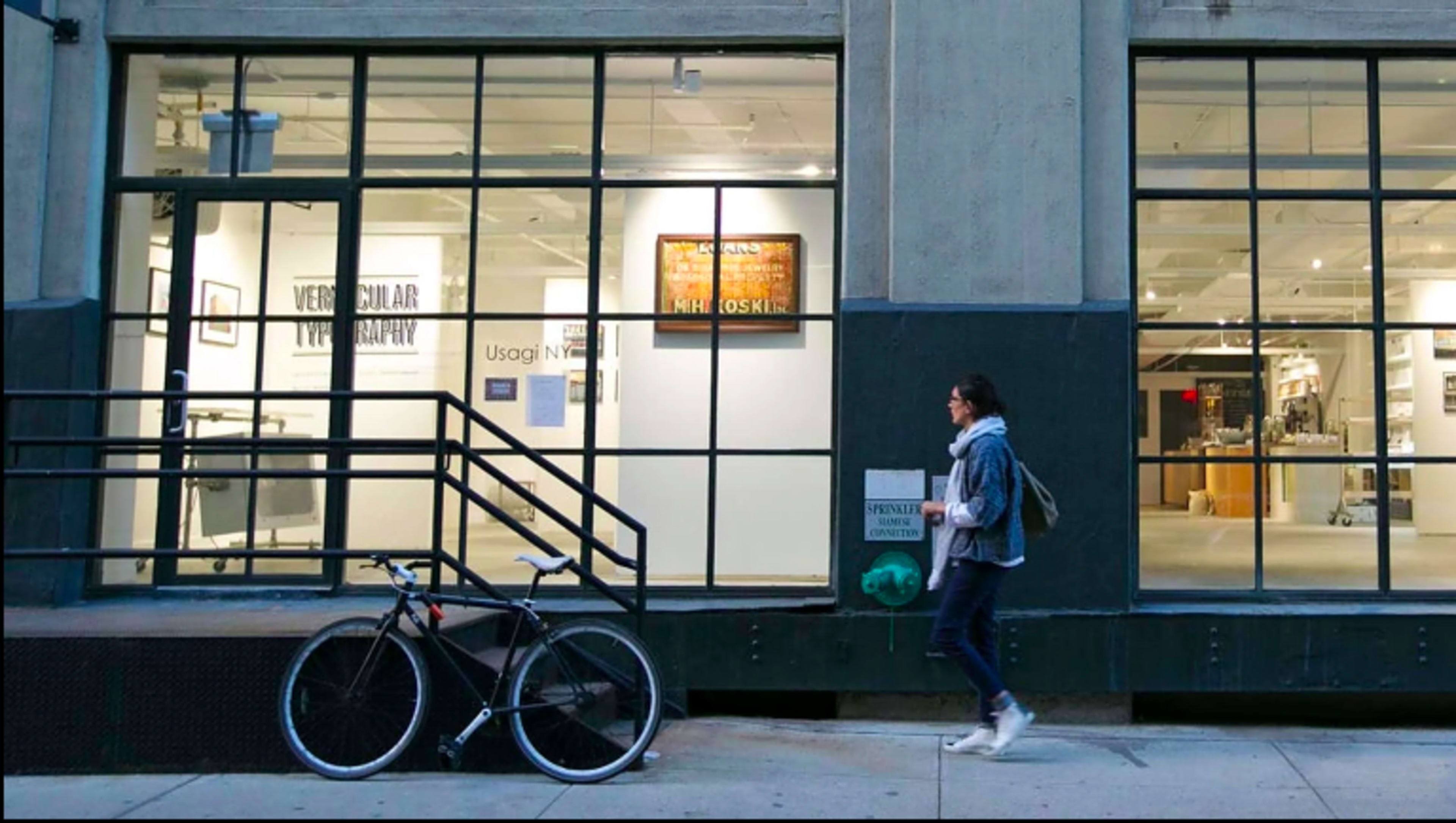 A woman walks past a storefront with large windows displaying artwork and a bicycle parked nearby.
