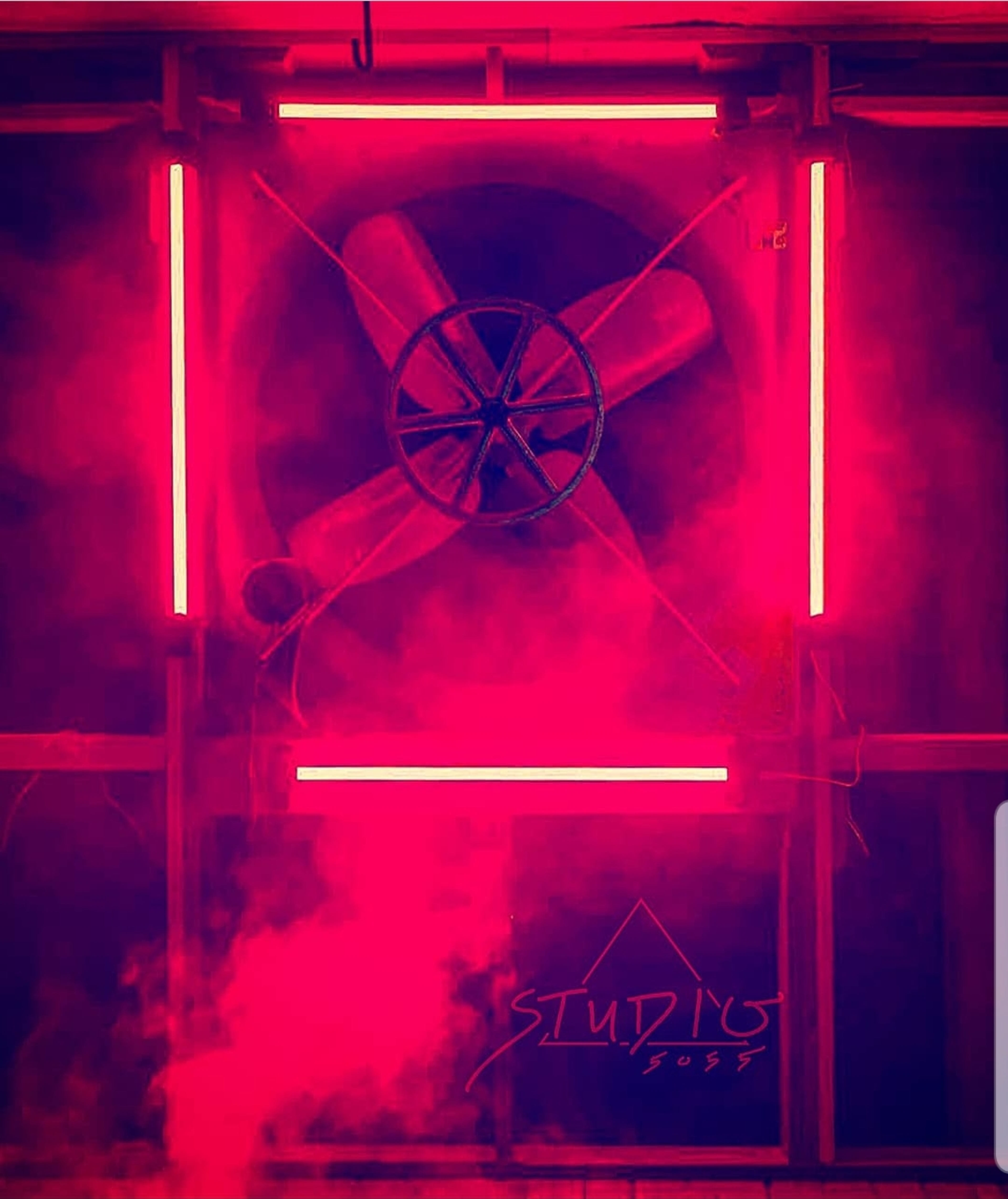 A large fan is framed by neon pink lights and surrounded by swirling fog.
