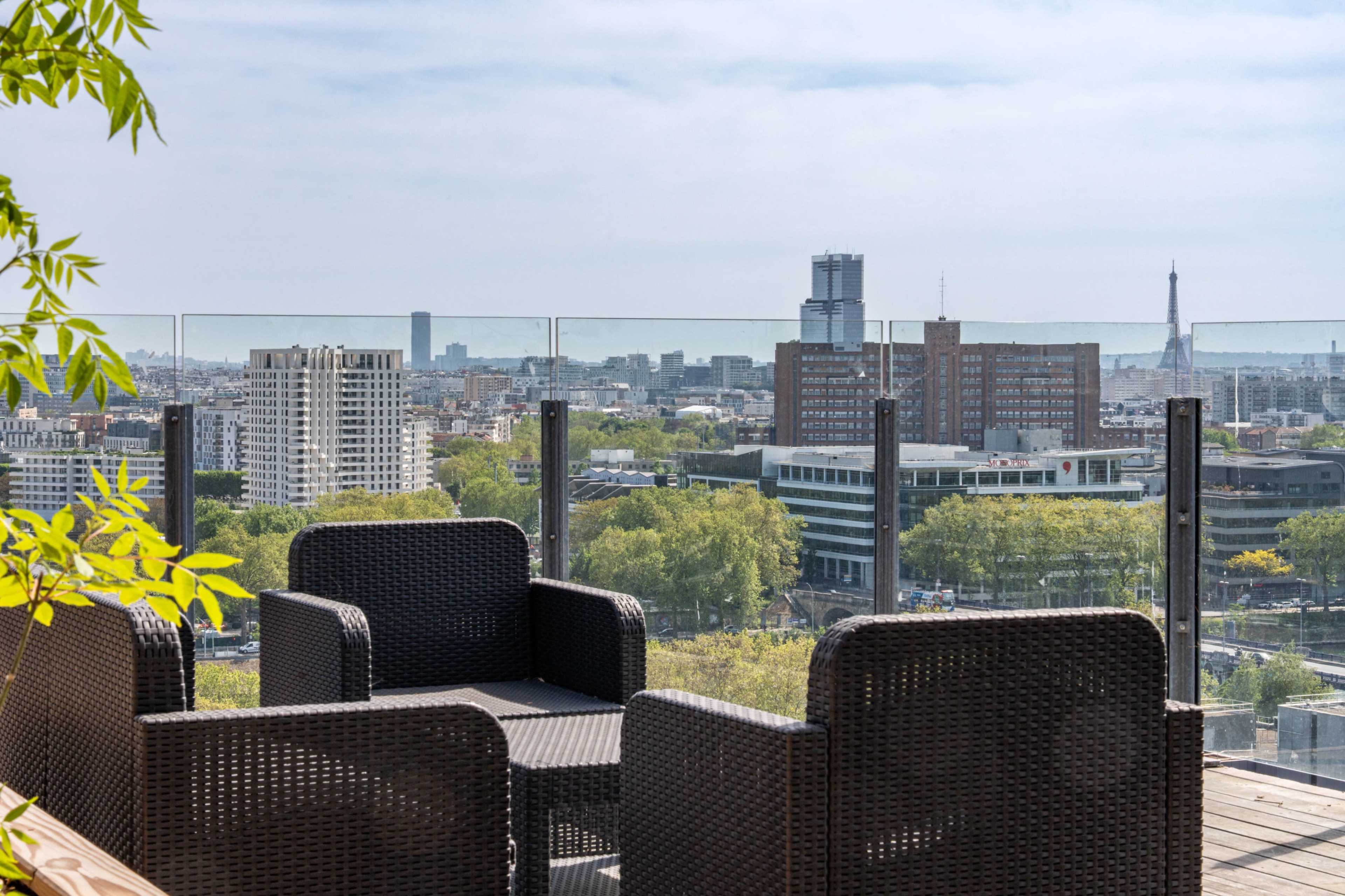 A rooftop terrace with wicker chairs overlooks a city skyline featuring modern buildings and the Eiffel Tower in the distance.