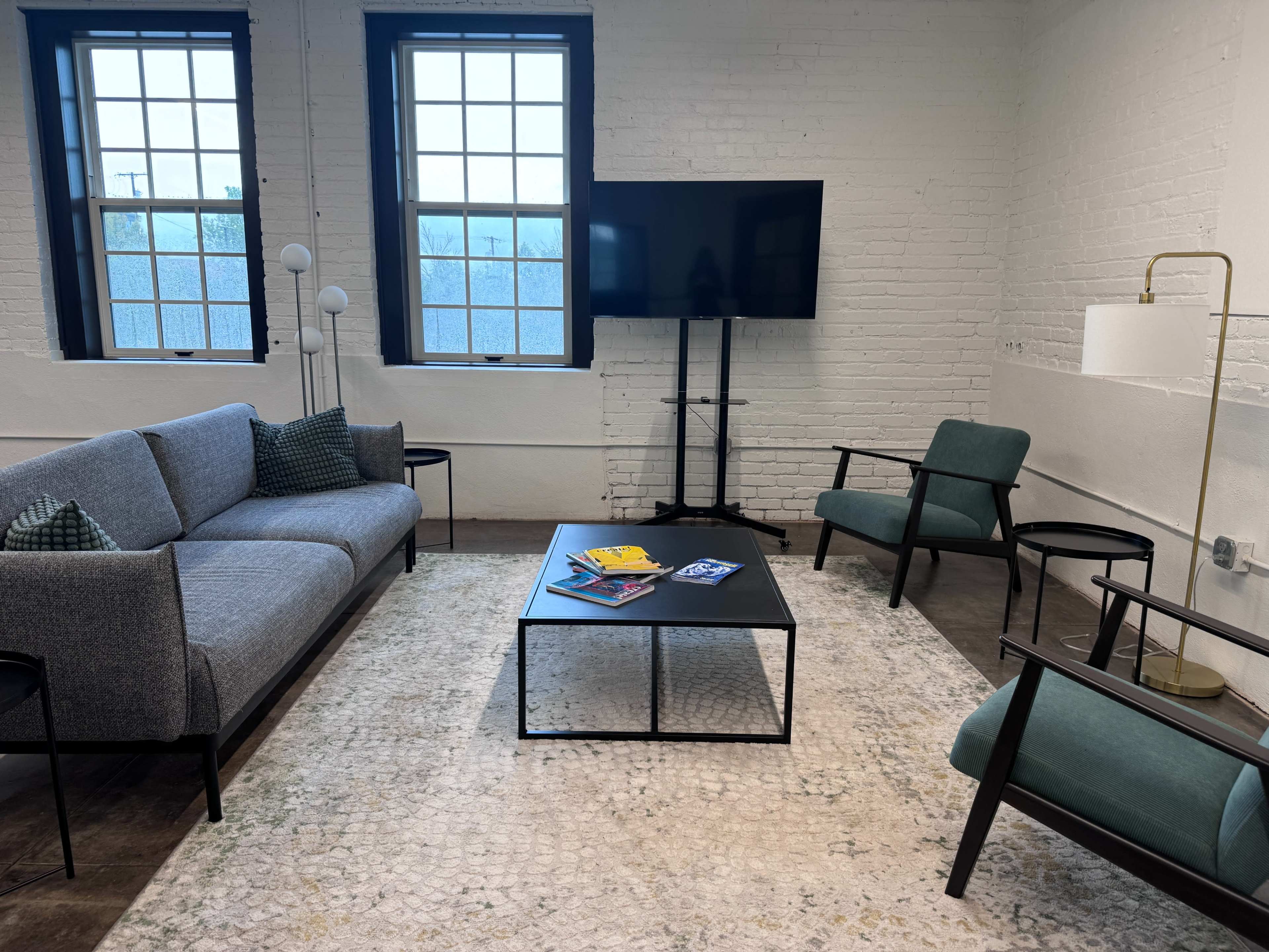 The image shows a modern lounge area with a gray sofa, two green chairs, a black coffee table, a television on a stand, and large windows allowing natural light.