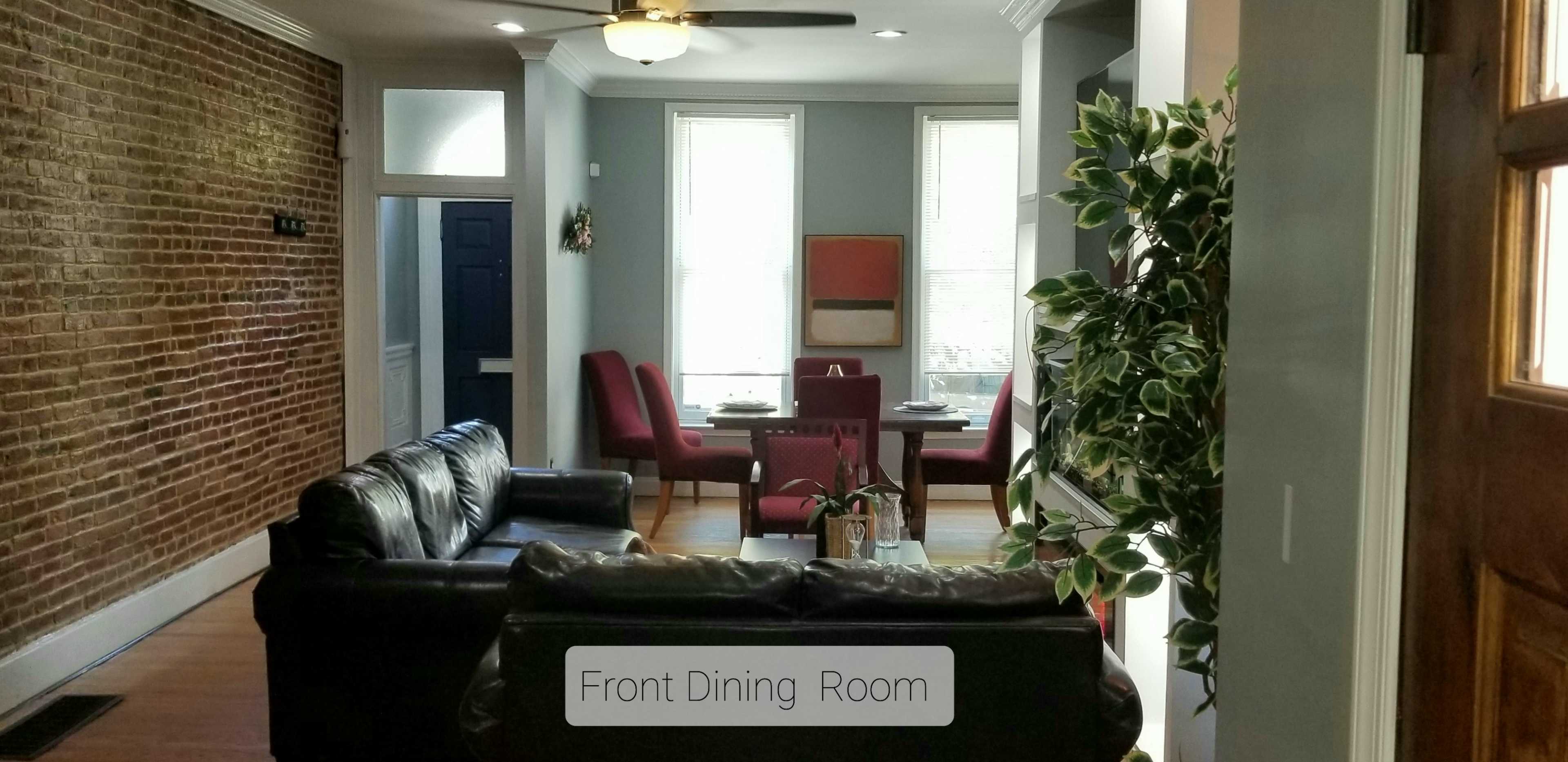 A living space with a brown leather sofa in the foreground and a dining area featuring a table set for four in the background, accompanied by exposed brick walls and large windows.