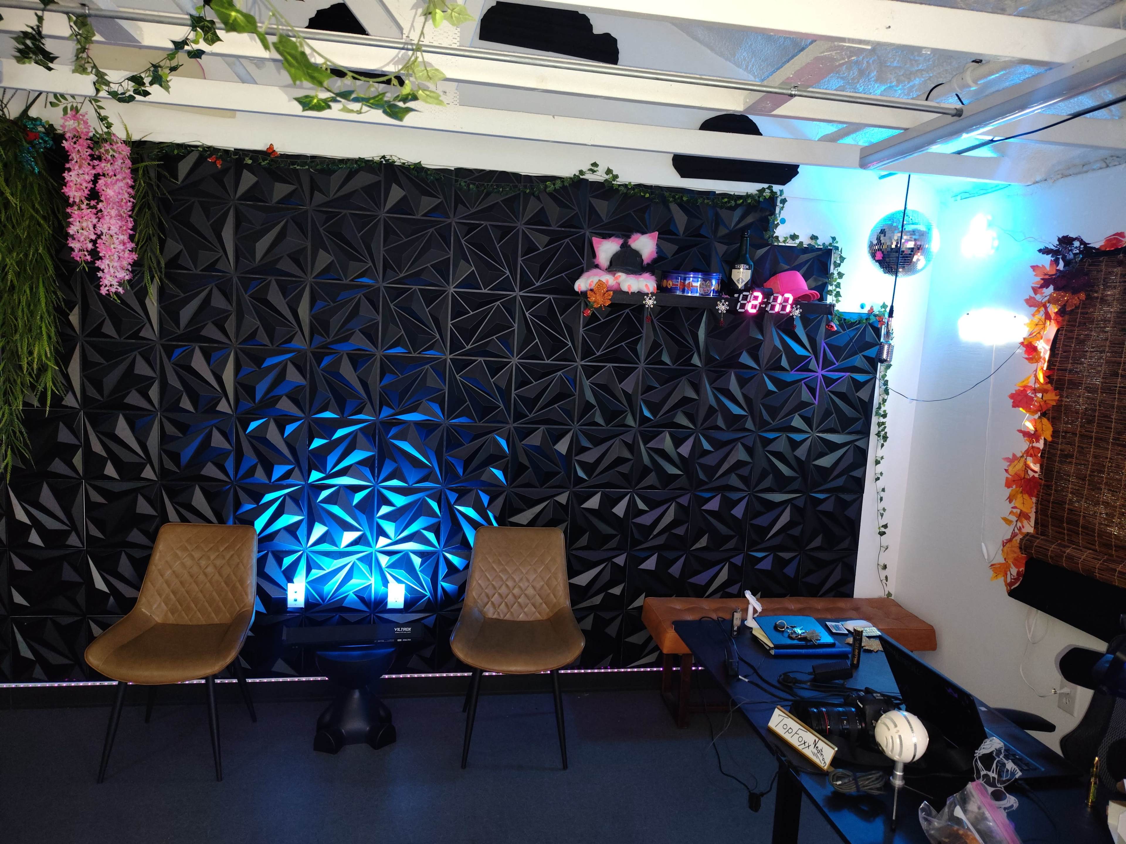 The scene features a modern room with a textured black wall, two beige chairs, a small black table, and various decorative elements like plants, lights, and wall shelves.