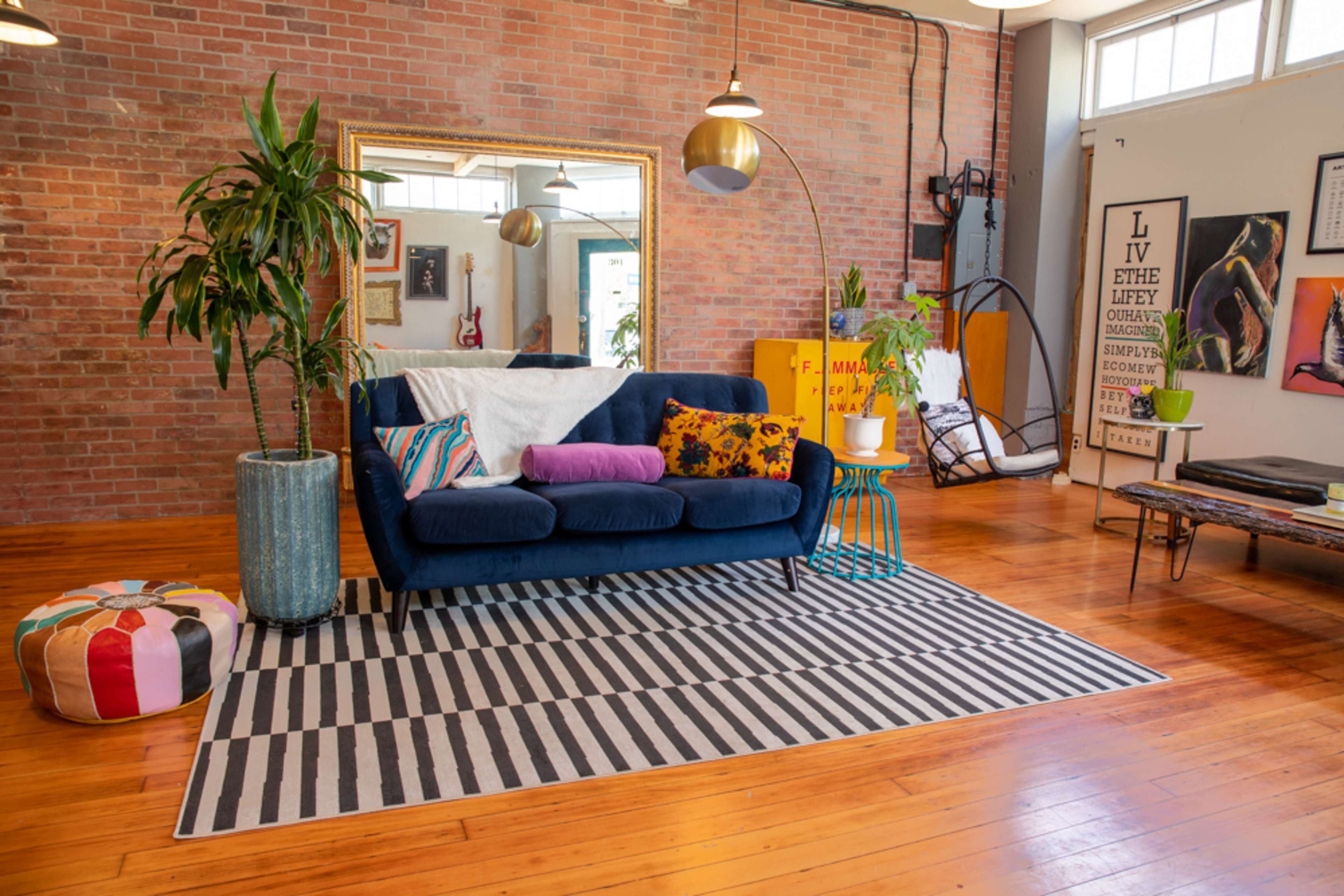 A bright living room features a blue sofa with colorful cushions, a large mirror, a potted plant, and a striped rug on polished wooden flooring.