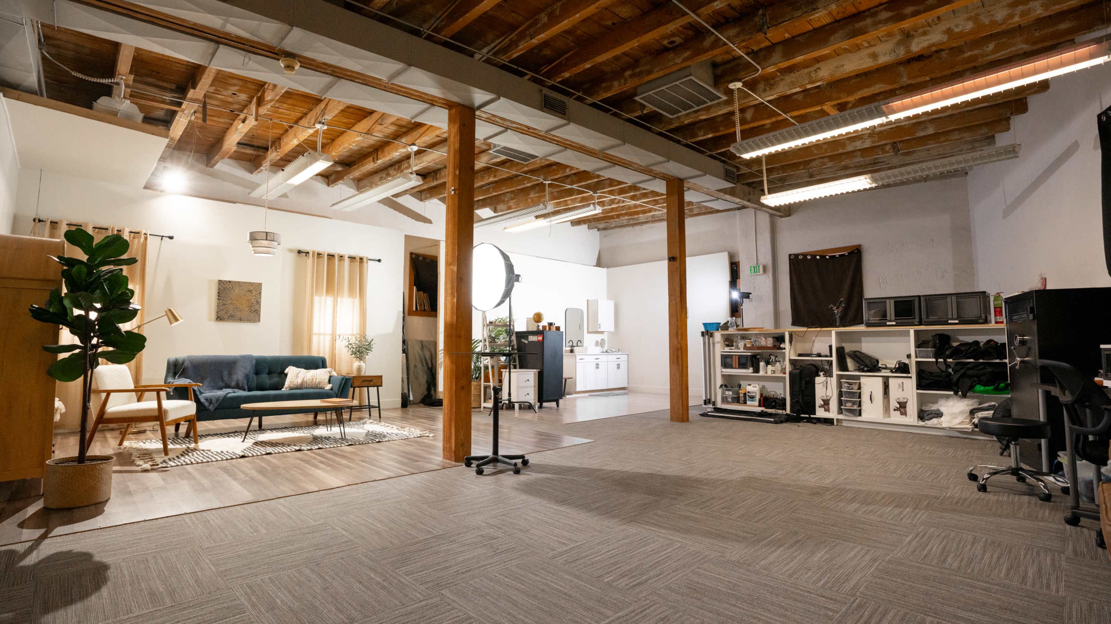 The image features a spacious, industrial-style interior with wooden beams, a seating area, and various furniture pieces, including a kitchenette and storage units.