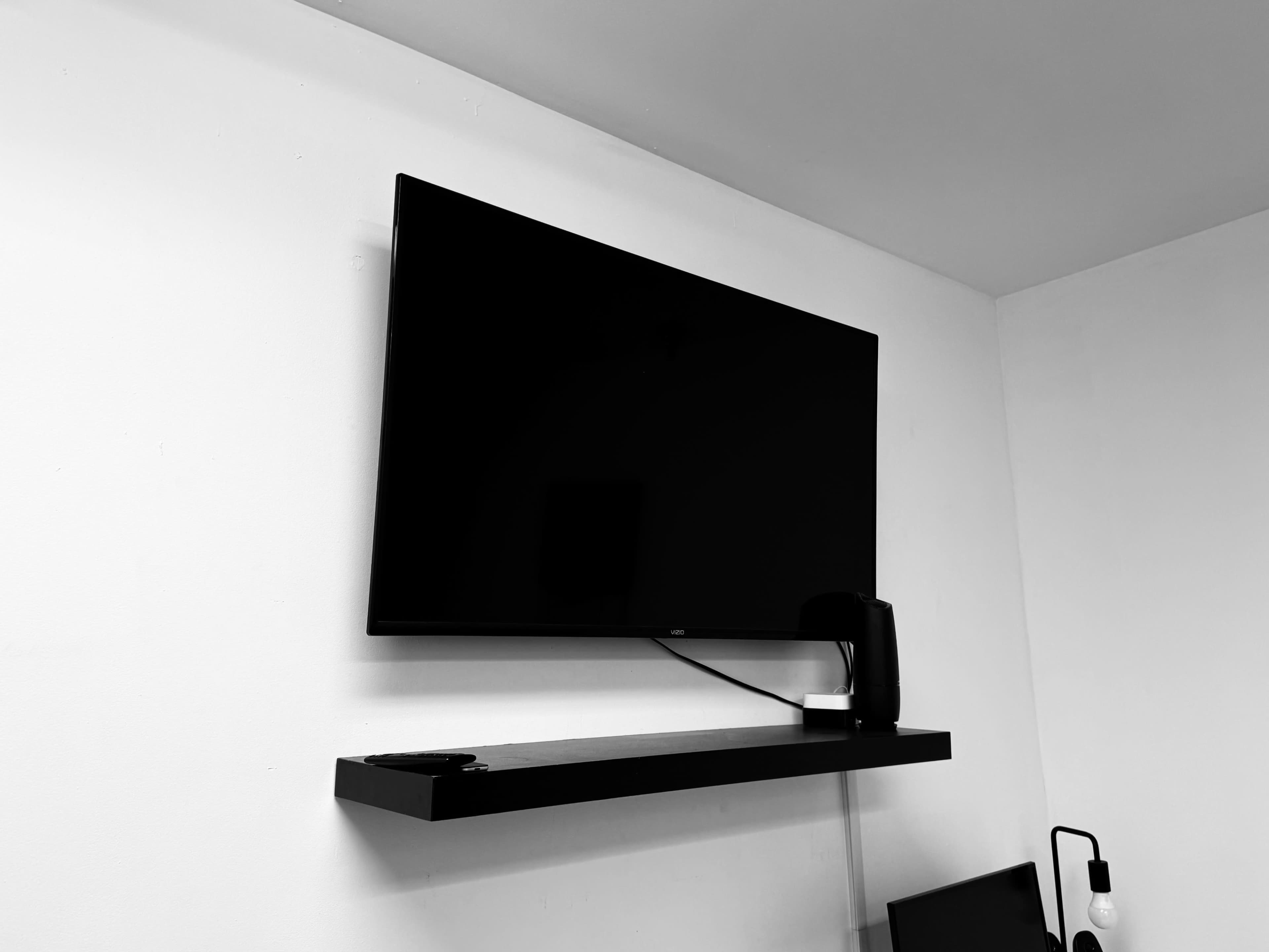 A black television is mounted on a white wall above a dark shelf.