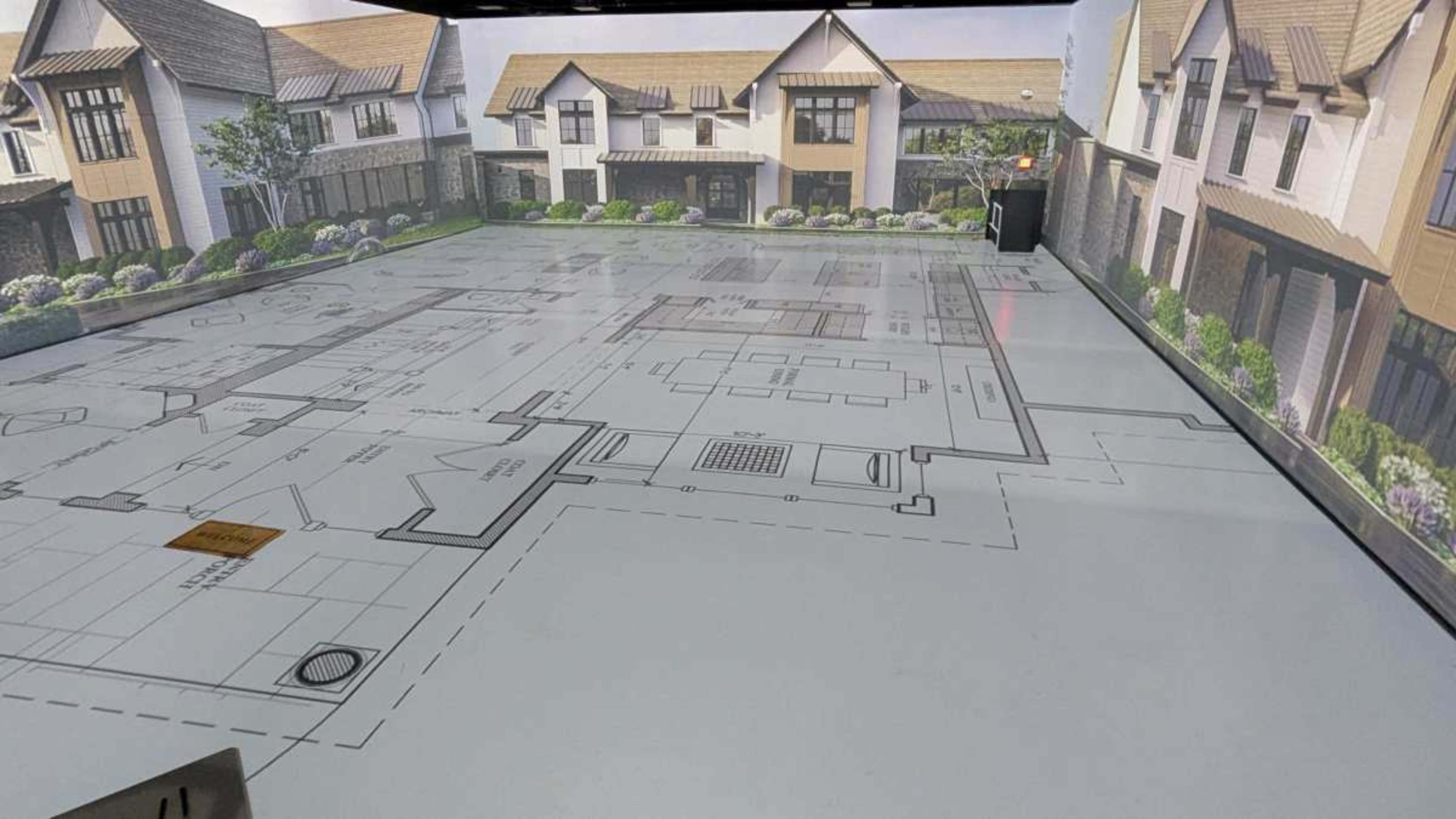 The image shows a floor space with a printed architectural blueprint of a house layout, surrounded by detailed visuals of exterior house designs and landscaping.
