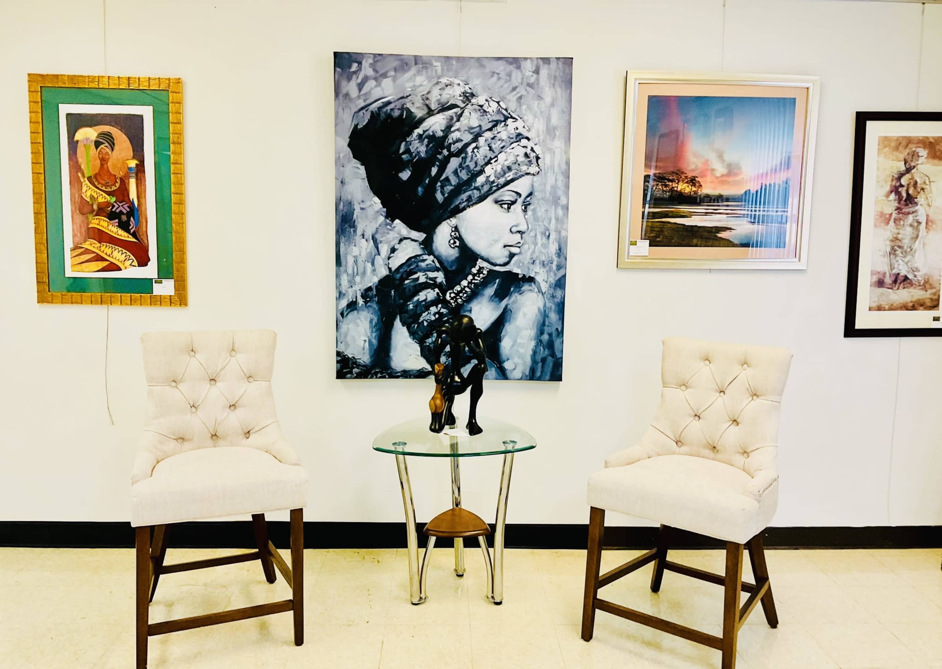 The image shows a small seating area with two upholstered chairs, a glass side table featuring a statue, and a diverse collection of framed artwork on the wall.