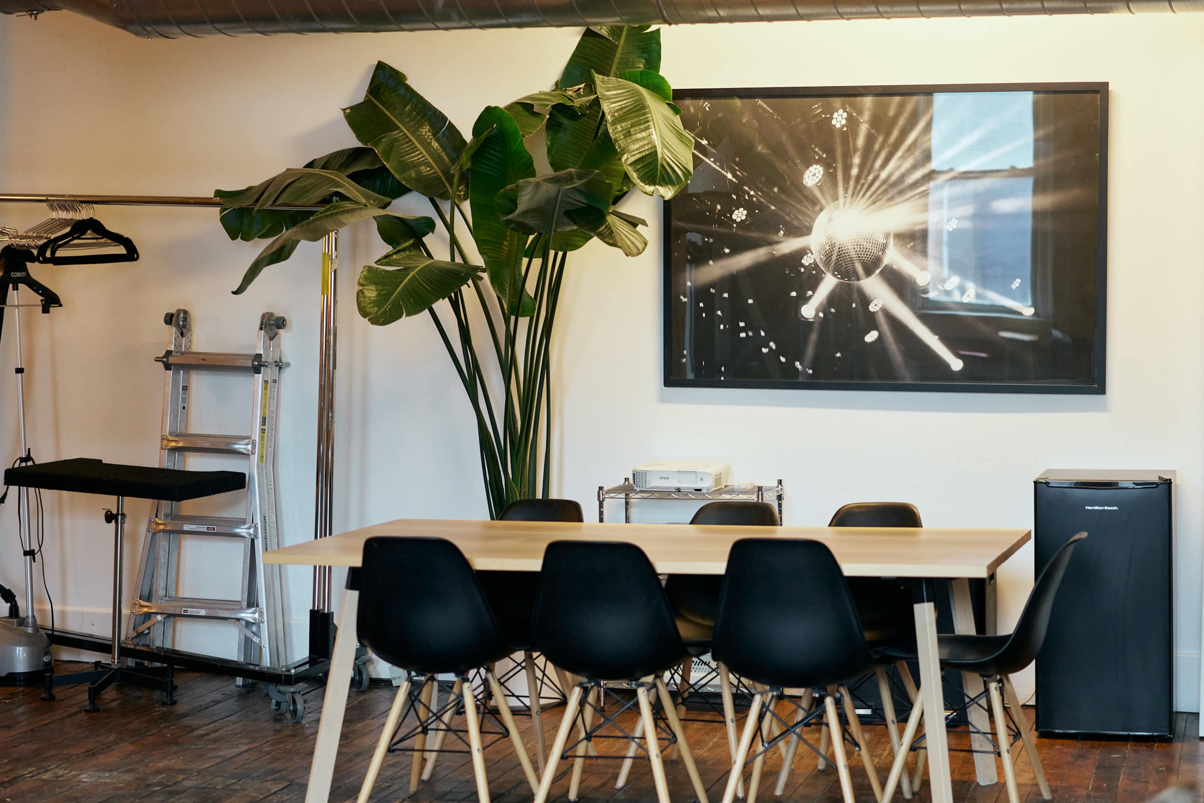 The image shows a modern meeting room featuring a large wooden table surrounded by black chairs, a tall plant, and a prominent wall art piece depicting a disco ball.