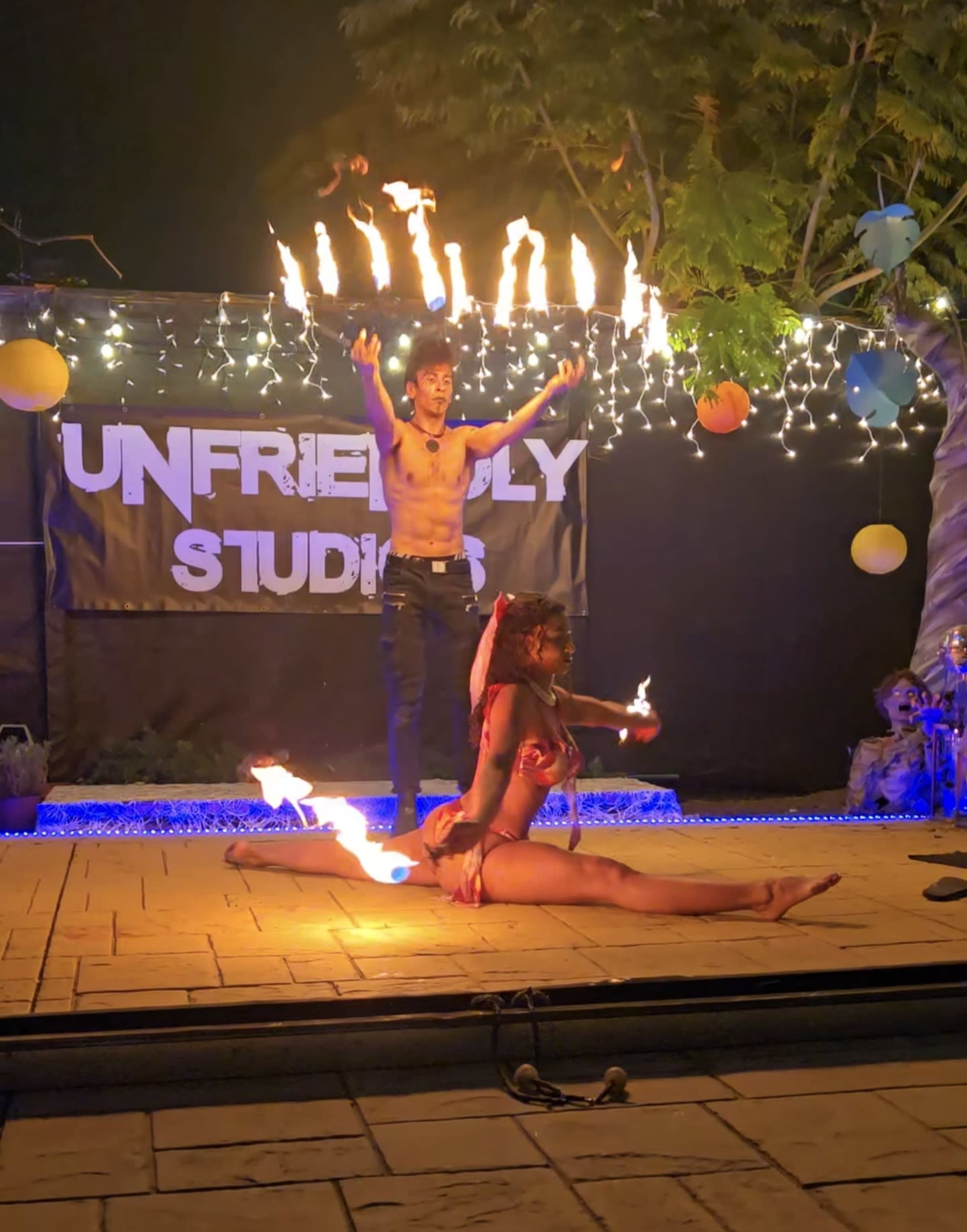 A fire performer dances with flaming props while another performer poses on the ground, both illuminated by colorful lights at an outdoor event.