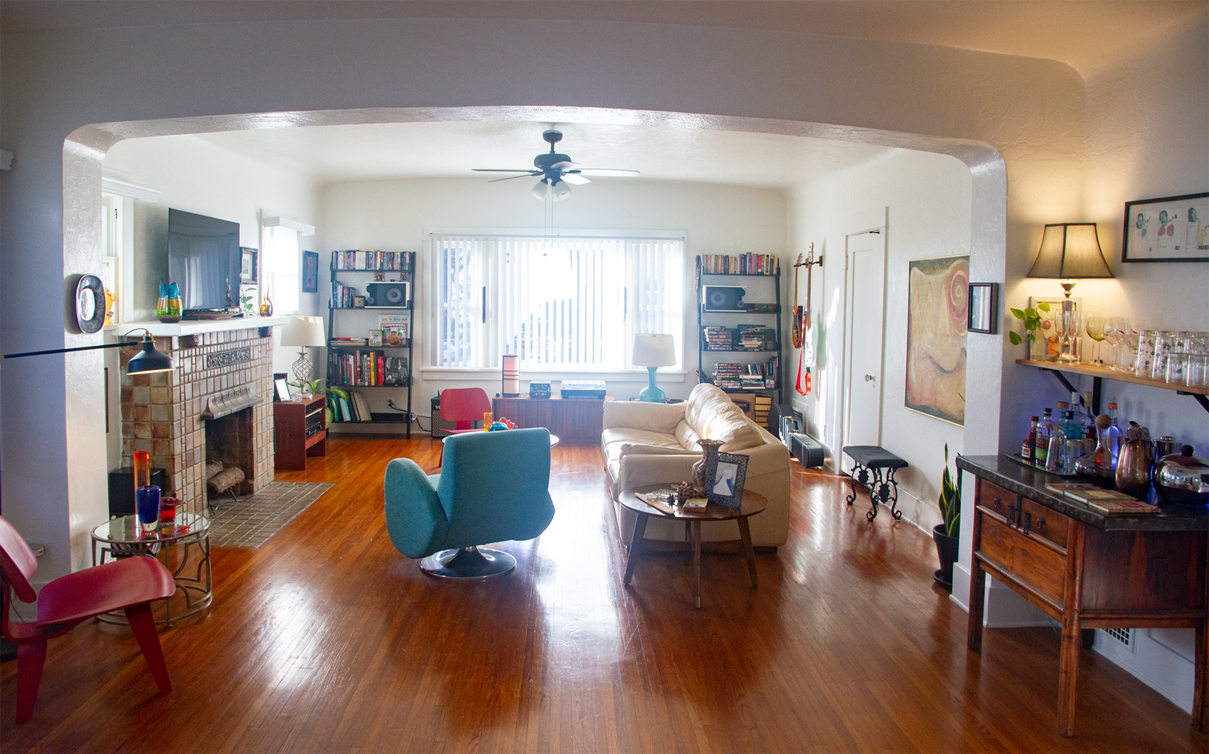 The living room features a fireplace, bookshelves, and a mix of seating arrangements, including a blue chair and a beige sofa, with hardwood floors and a view of windows in the background.