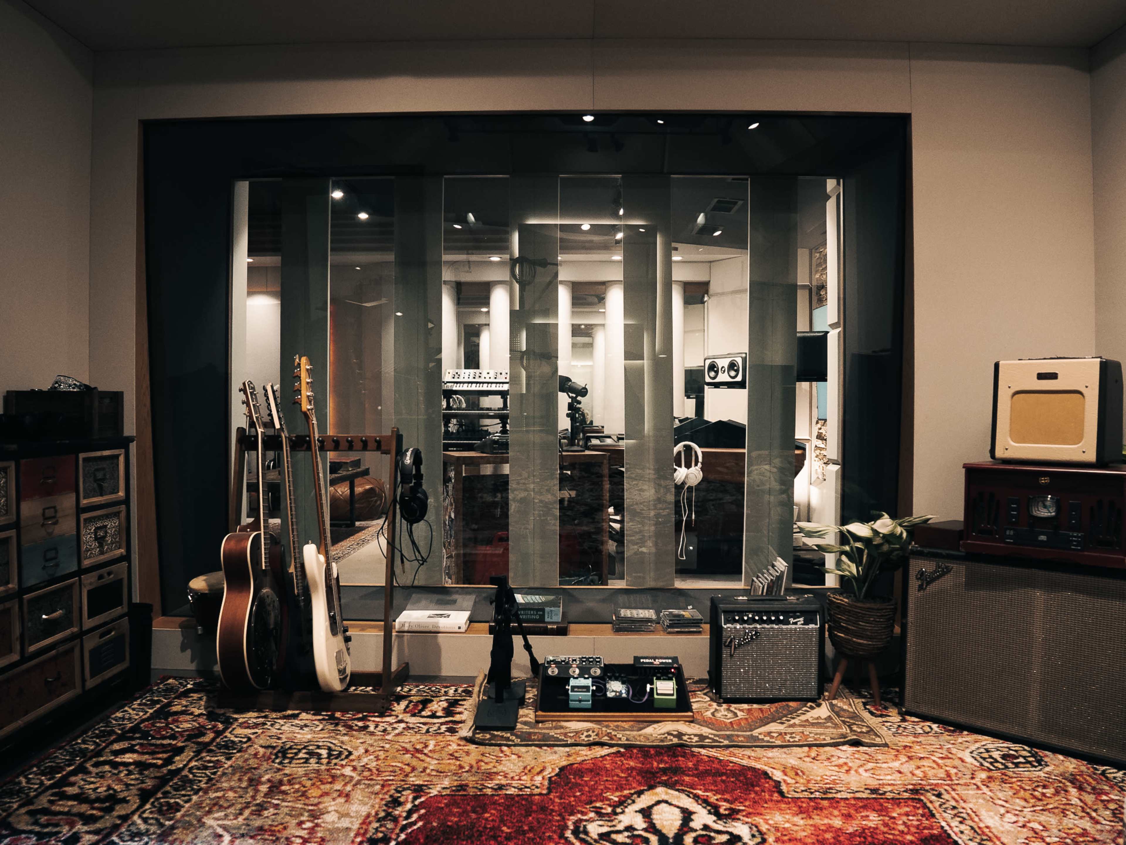 A music recording studio features guitars, amplifiers, and effect pedals arranged around a glass-walled recording area with a patterned rug on the floor.