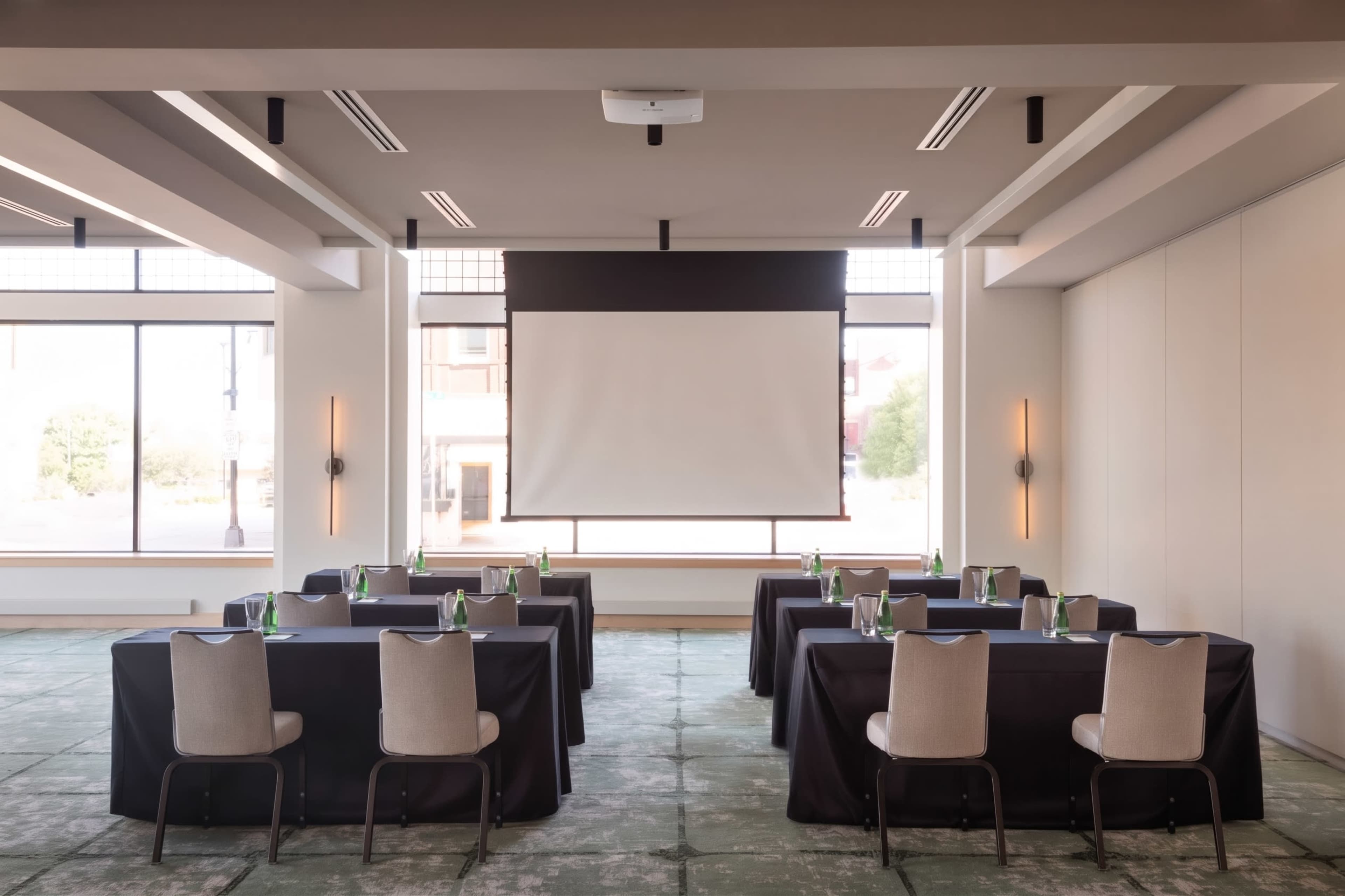 A conference room is set up with several tables covered in black cloth and chairs arranged for a meeting, with a large screen mounted on the wall at the front.