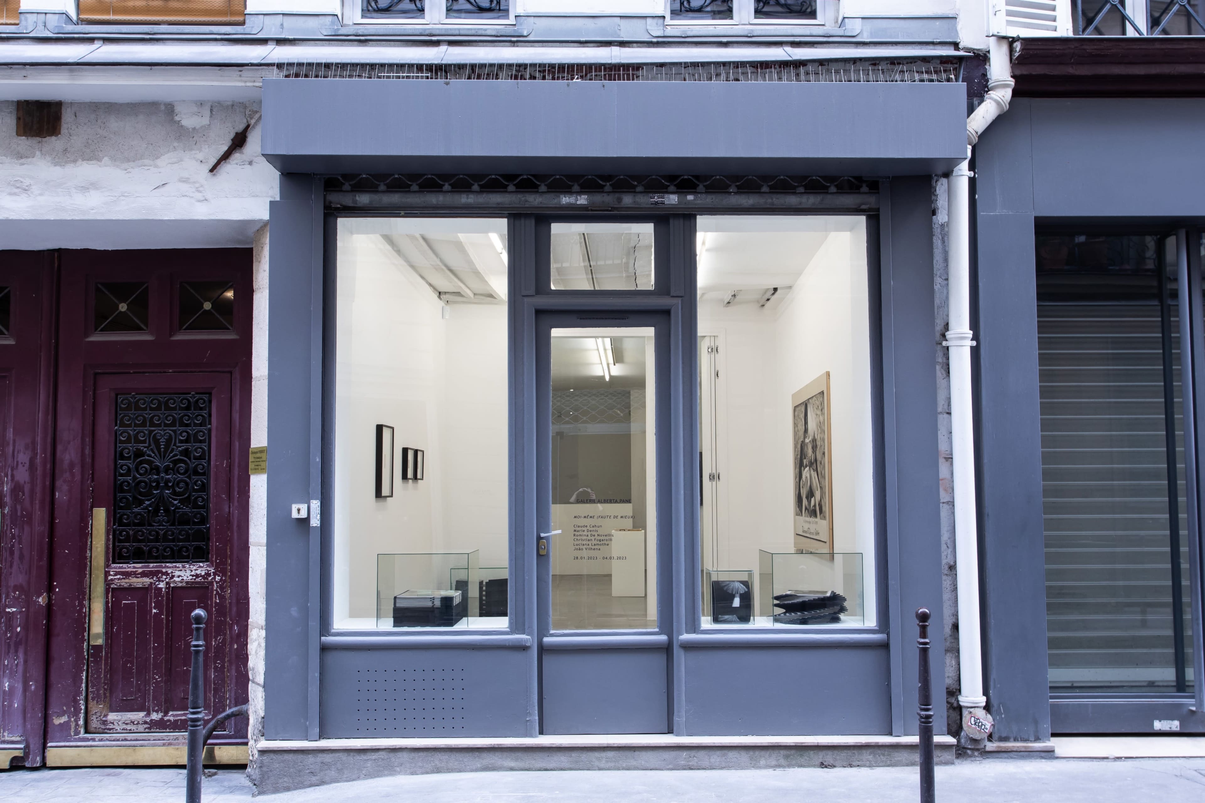 The image shows the exterior of a modern art gallery with large glass windows and a gray facade, flanked by a red door and a storefront with closed shutters.