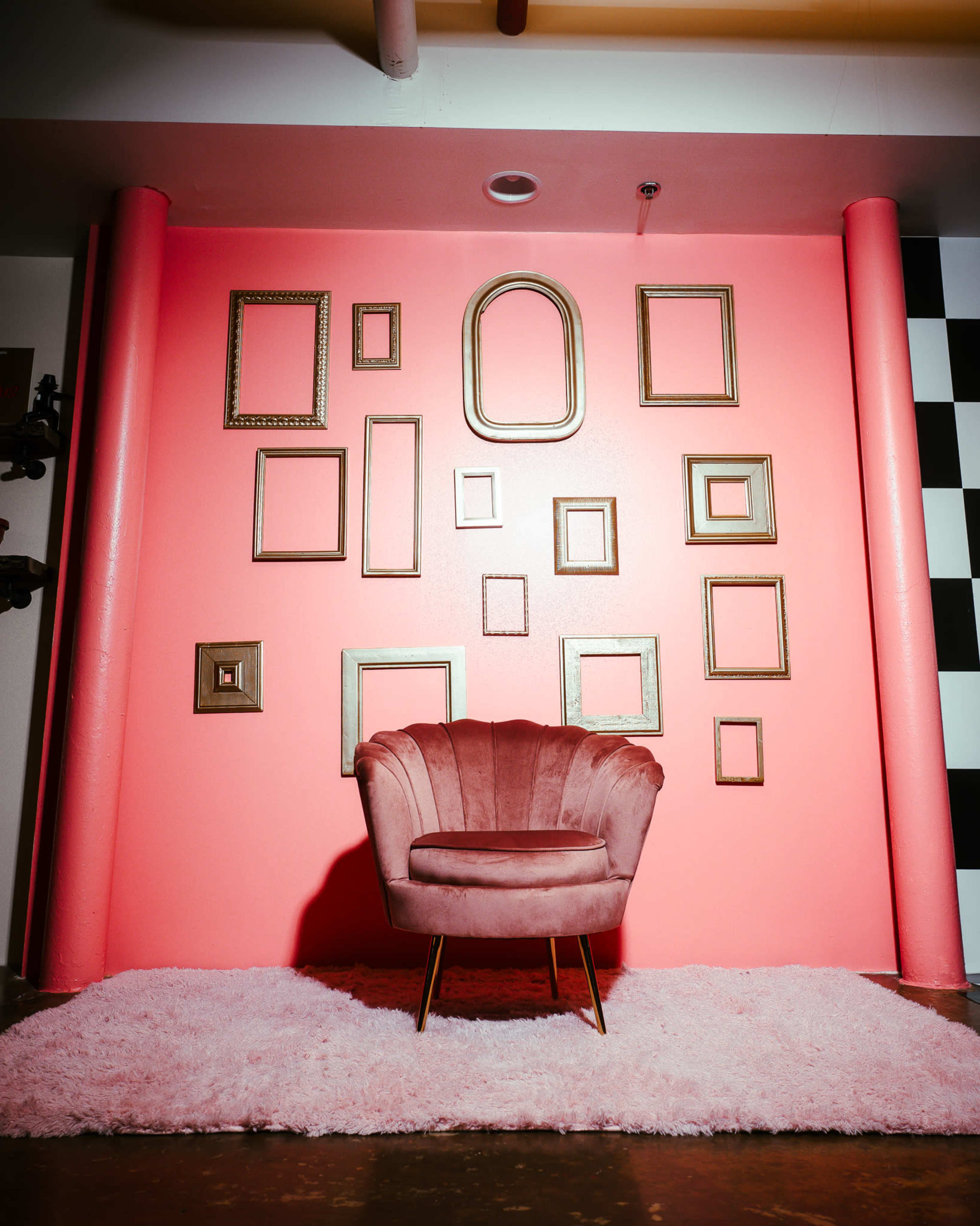 A plush pink chair on a soft rug in front of a vibrant pink wall adorned with various empty gold picture frames.