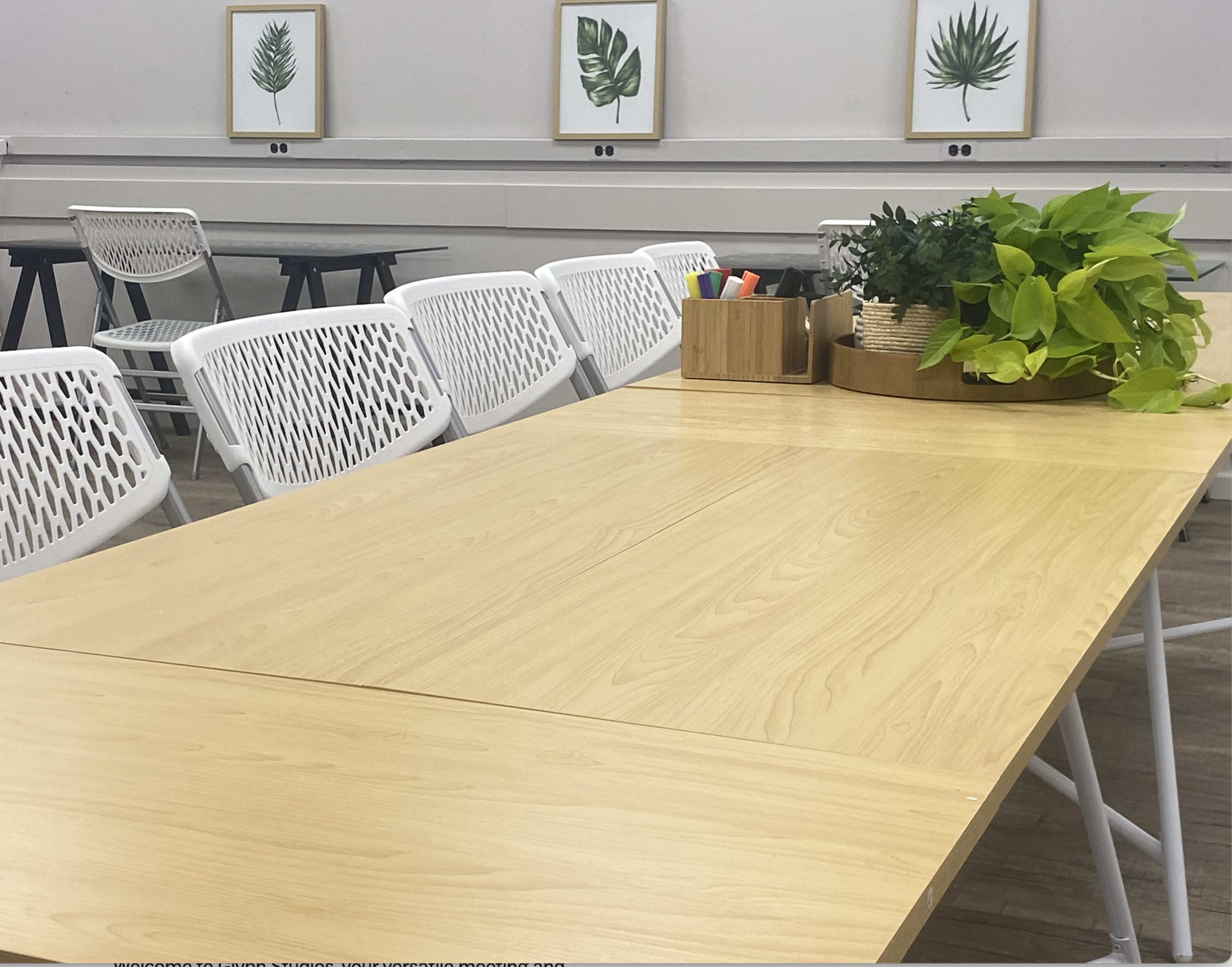 A light wooden table with a potted plant and stationery organizer is positioned in a room featuring framed leaf prints on the walls.