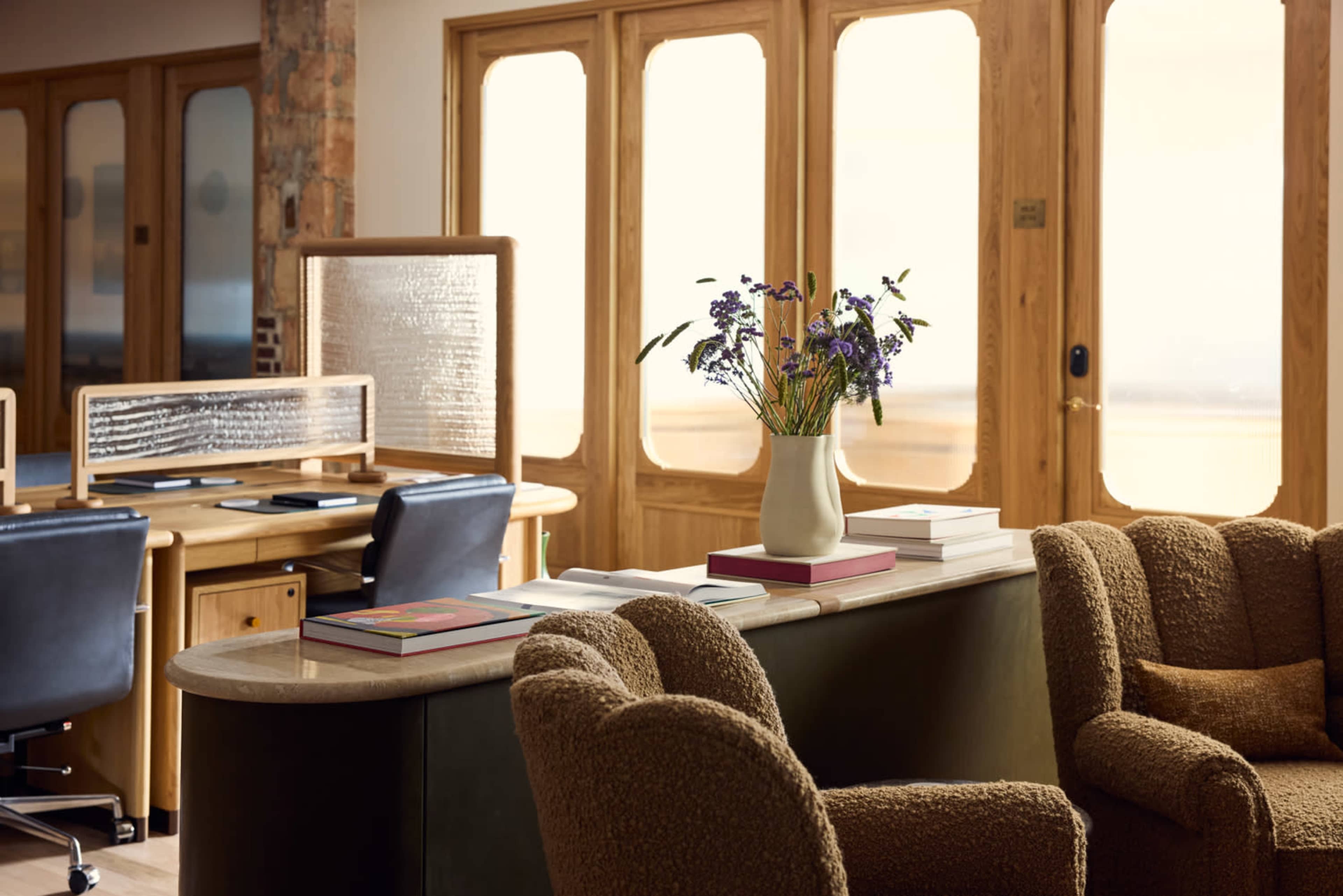 The image shows a stylish interior featuring a round table with books and a vase of flowers, alongside a cozy seating area and wooden doors leading to a bright outdoor space.