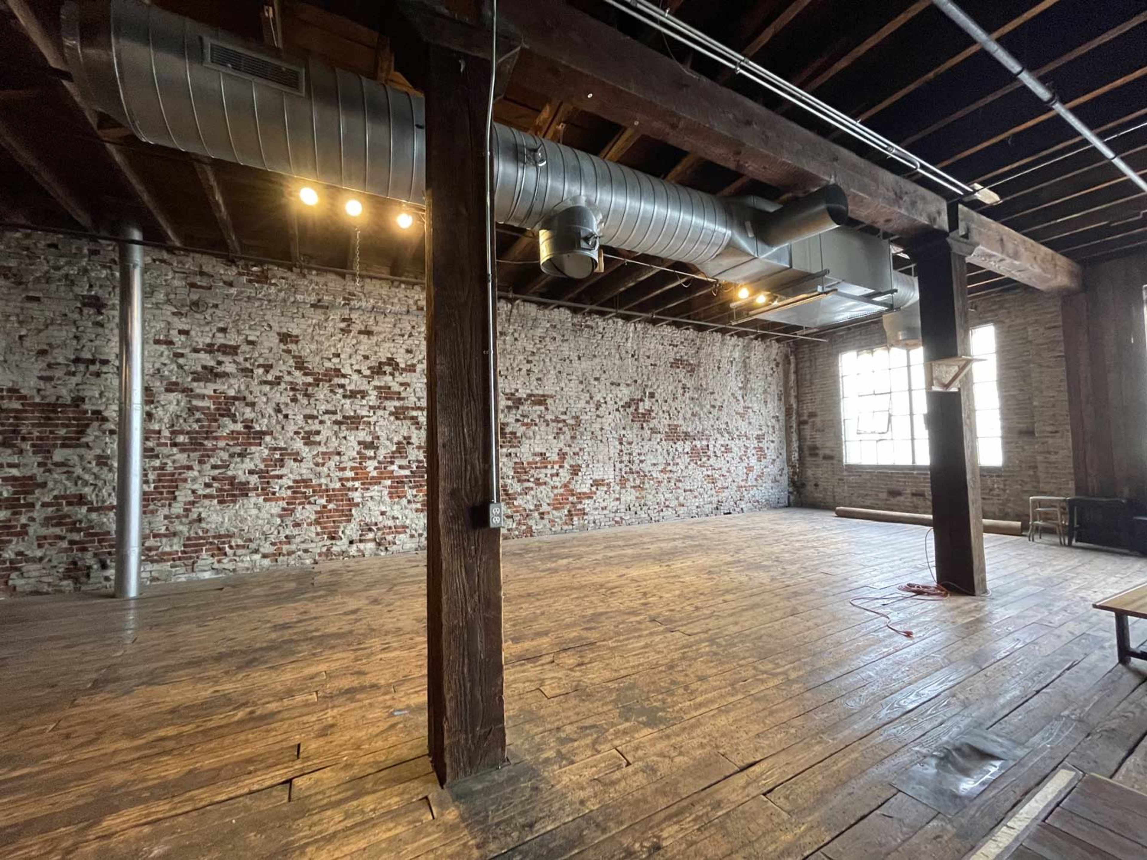 The image features an empty industrial space with wooden flooring, exposed brick walls, large windows, and visible ductwork along the ceiling.