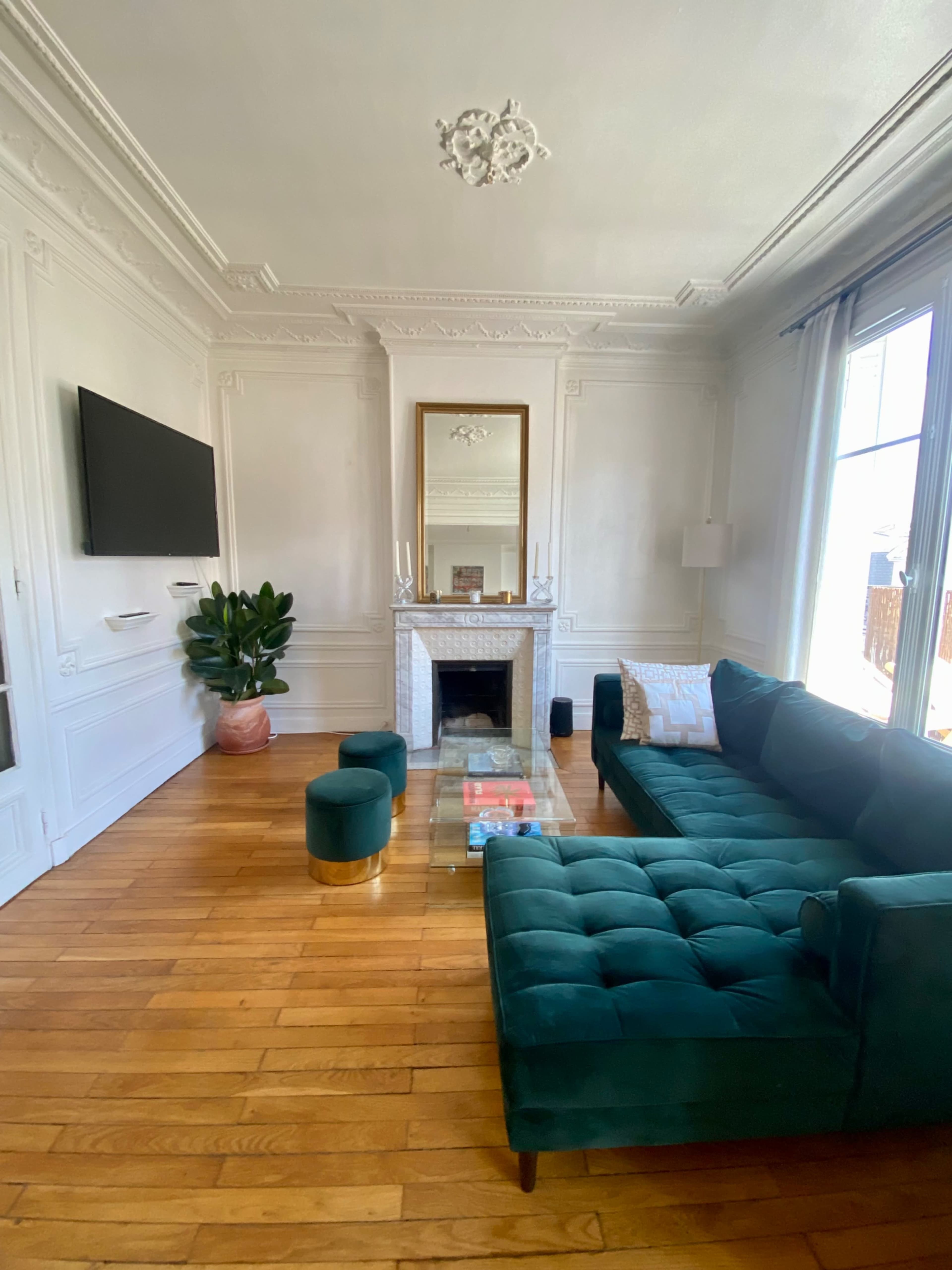 A stylish living room featuring a green velvet sectional sofa, a glass coffee table, a fireplace with a mirror above it, and a large flat-screen TV on the wall.