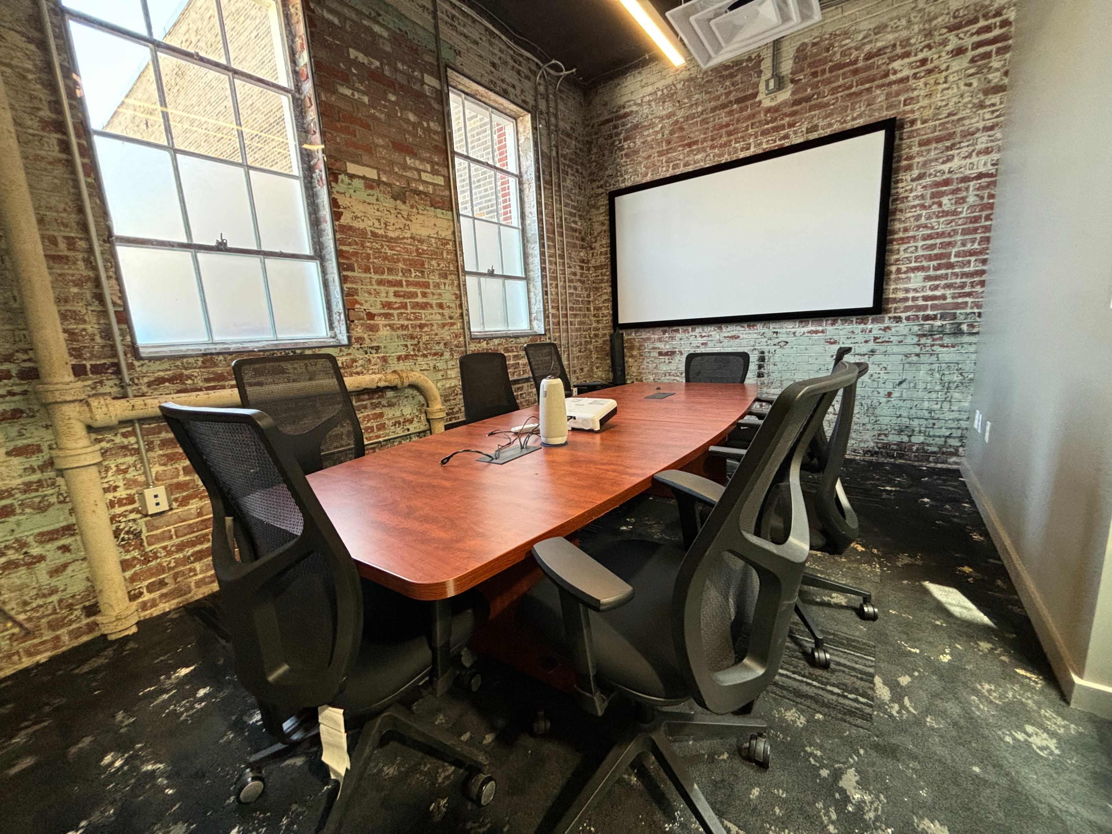 A conference room with a wooden table surrounded by office chairs, a projector, and brick walls.