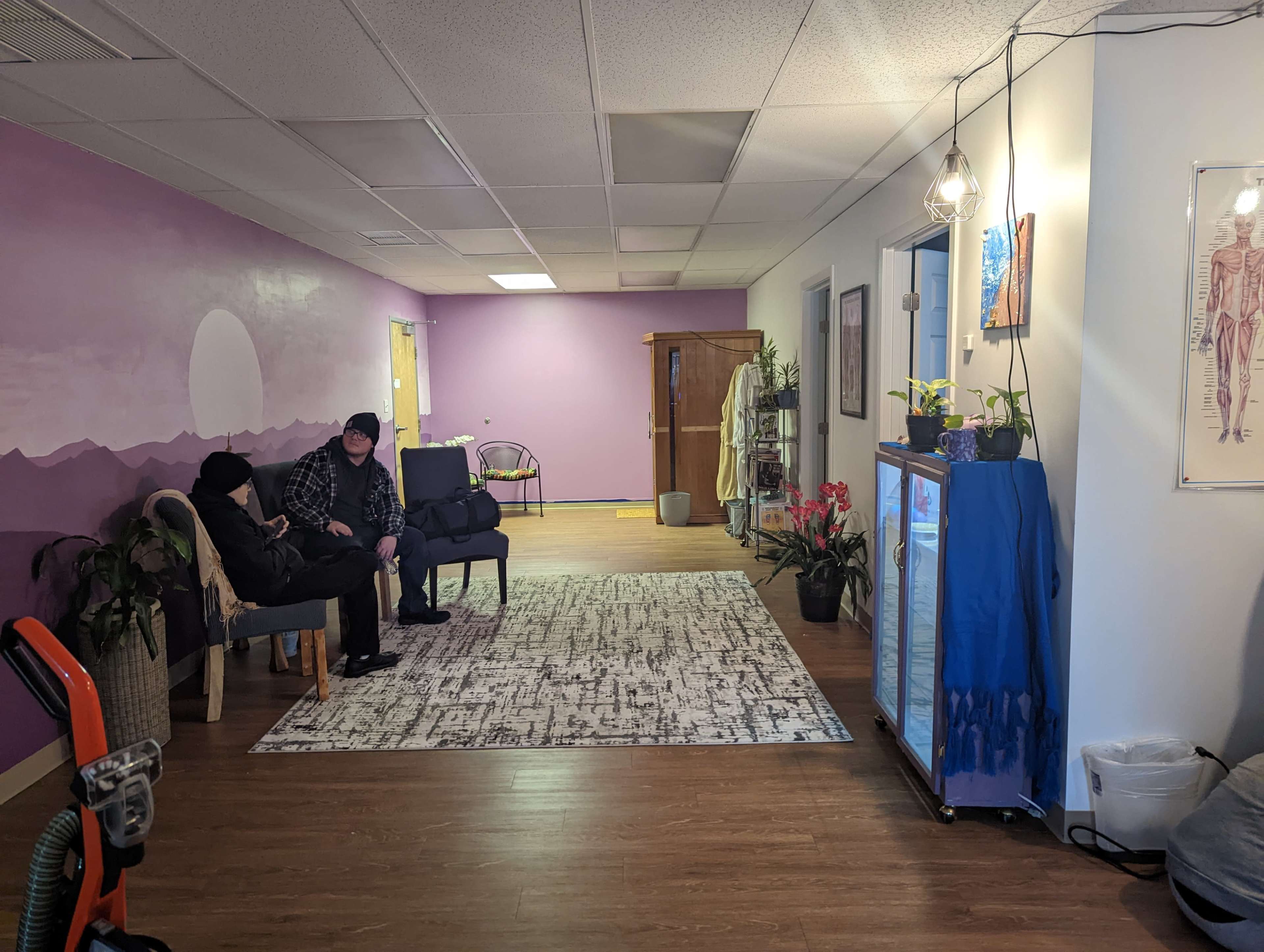 A waiting area with two people sitting on chairs, a purple wall mural in the background, and plants arranged throughout the space.