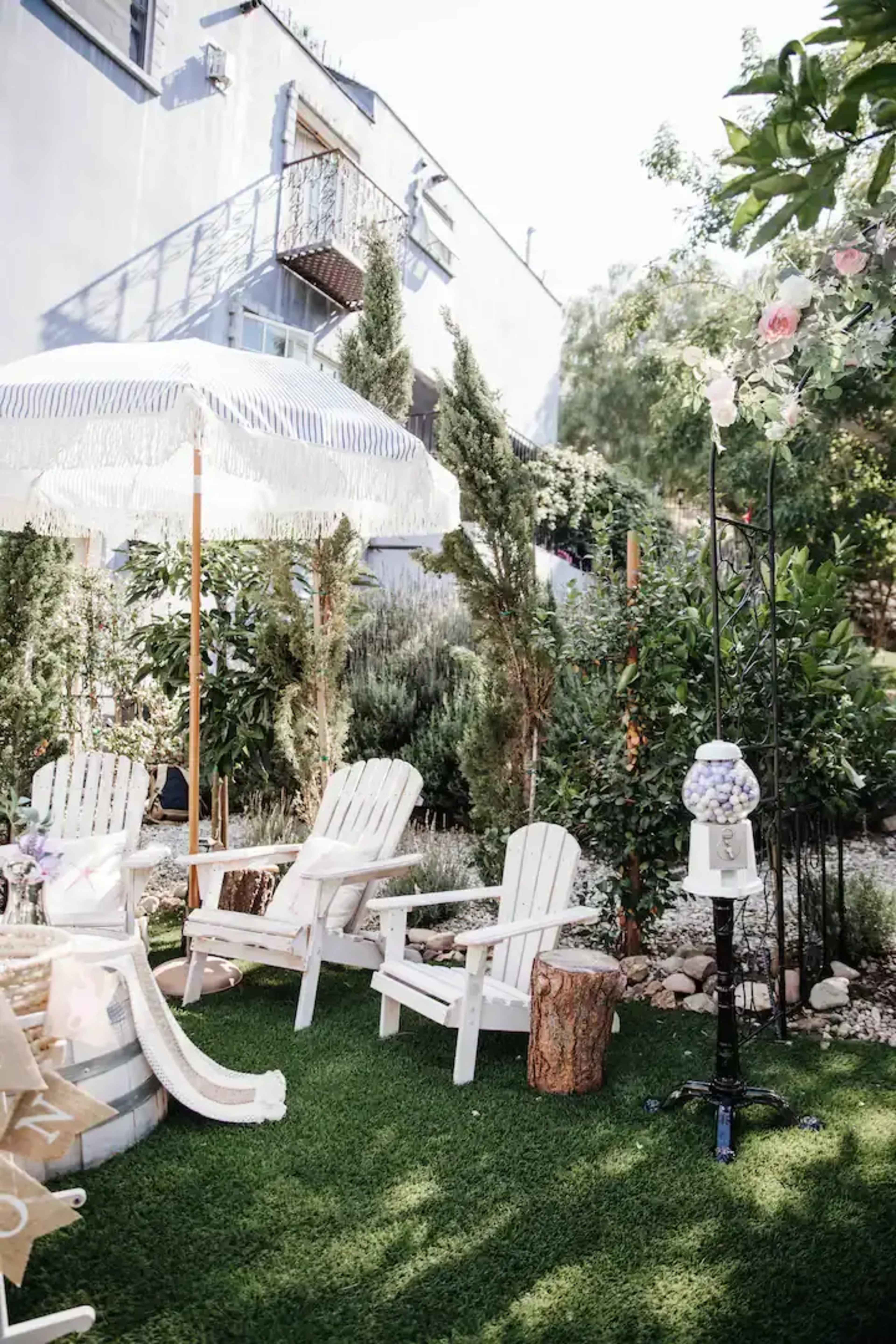 A garden area with white Adirondack chairs, a decorative umbrella, and a gumball machine on a grassy surface surrounded by lush greenery.