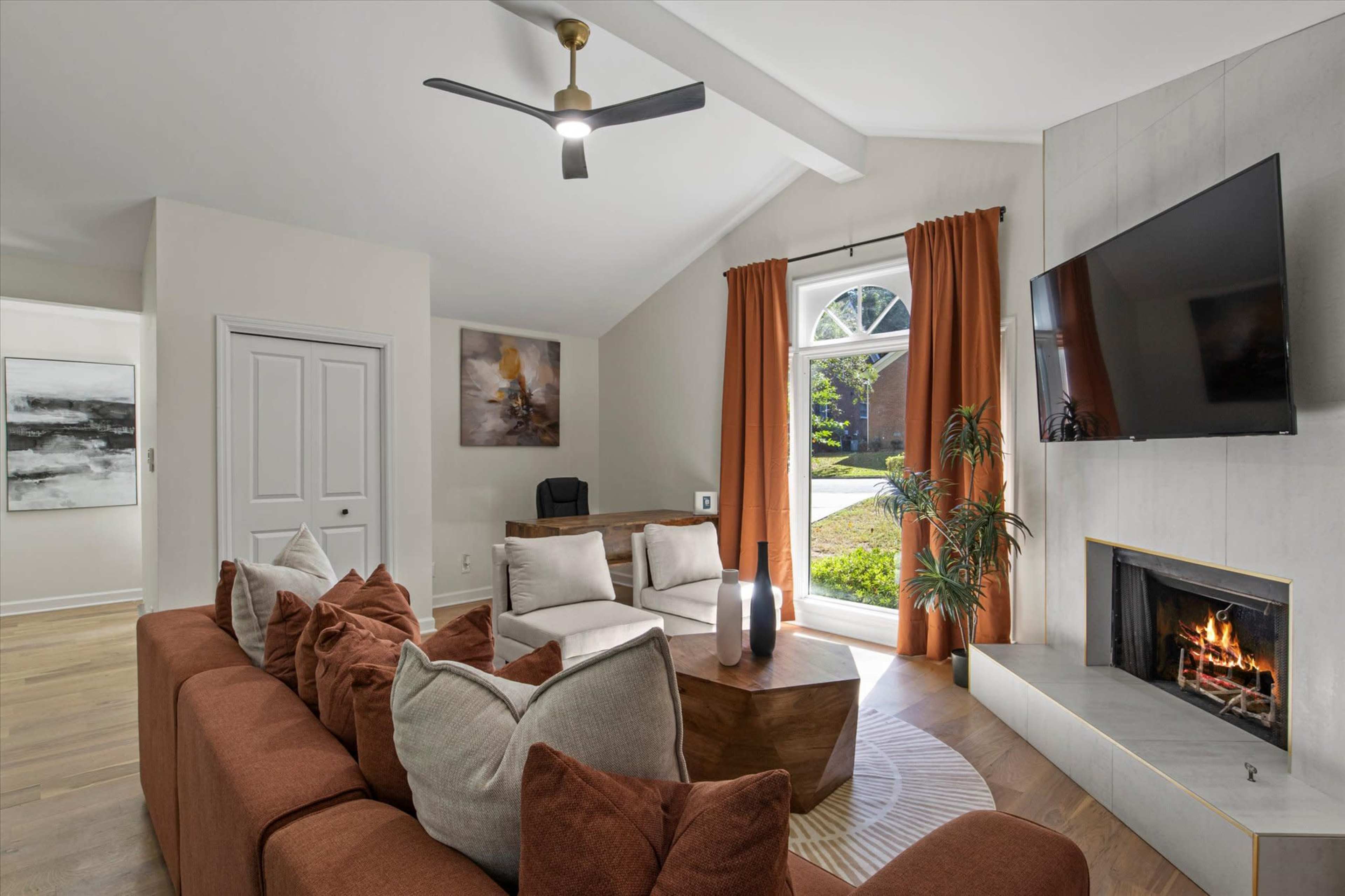 The image shows a modern living room with a brown sectional sofa, a wooden coffee table, and a fireplace, all under a sloped ceiling with a large window and a ceiling fan.