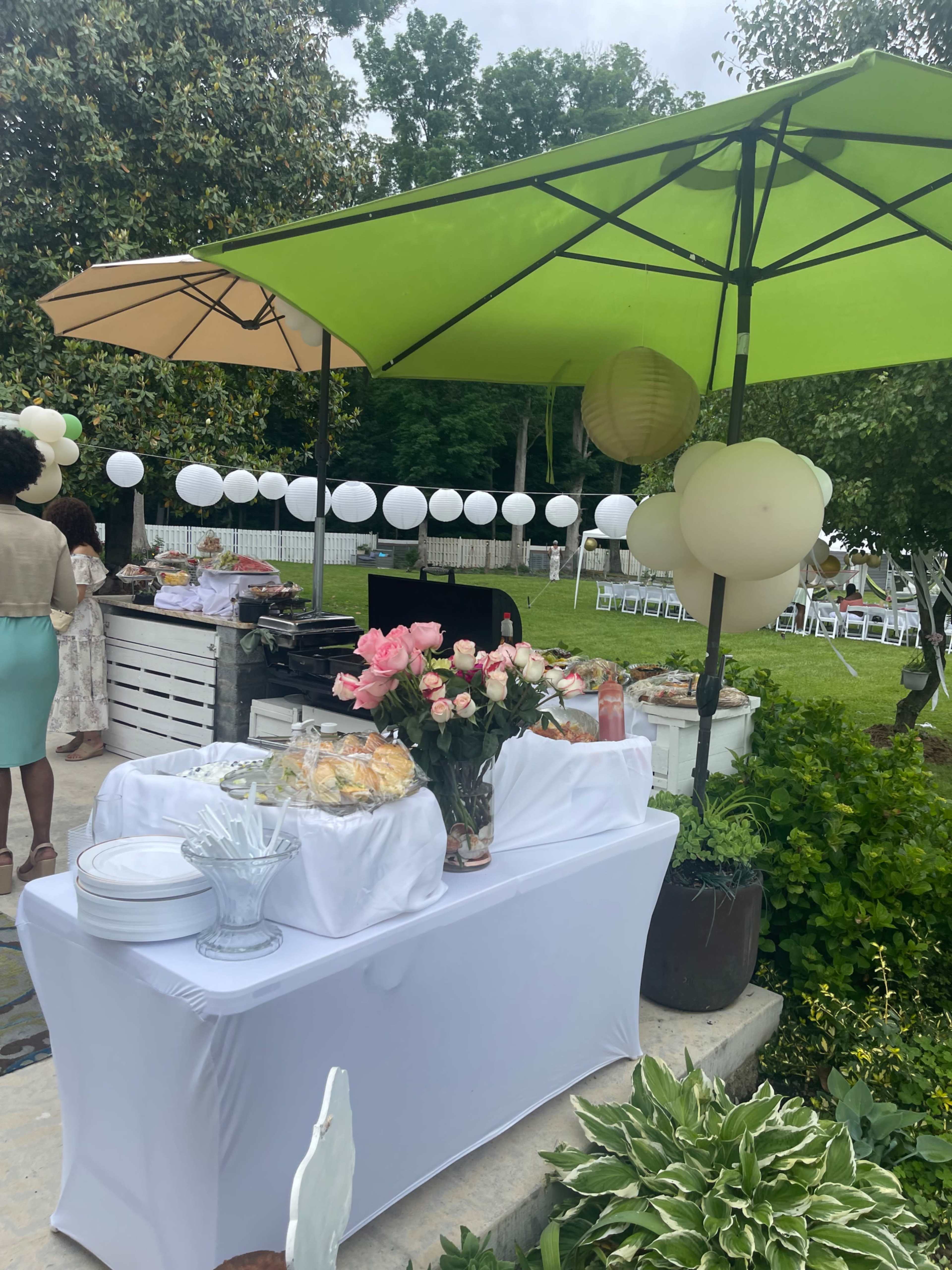 A buffet table with food and floral arrangements is set up outdoors under green and beige umbrellas, with white decorations in the background.