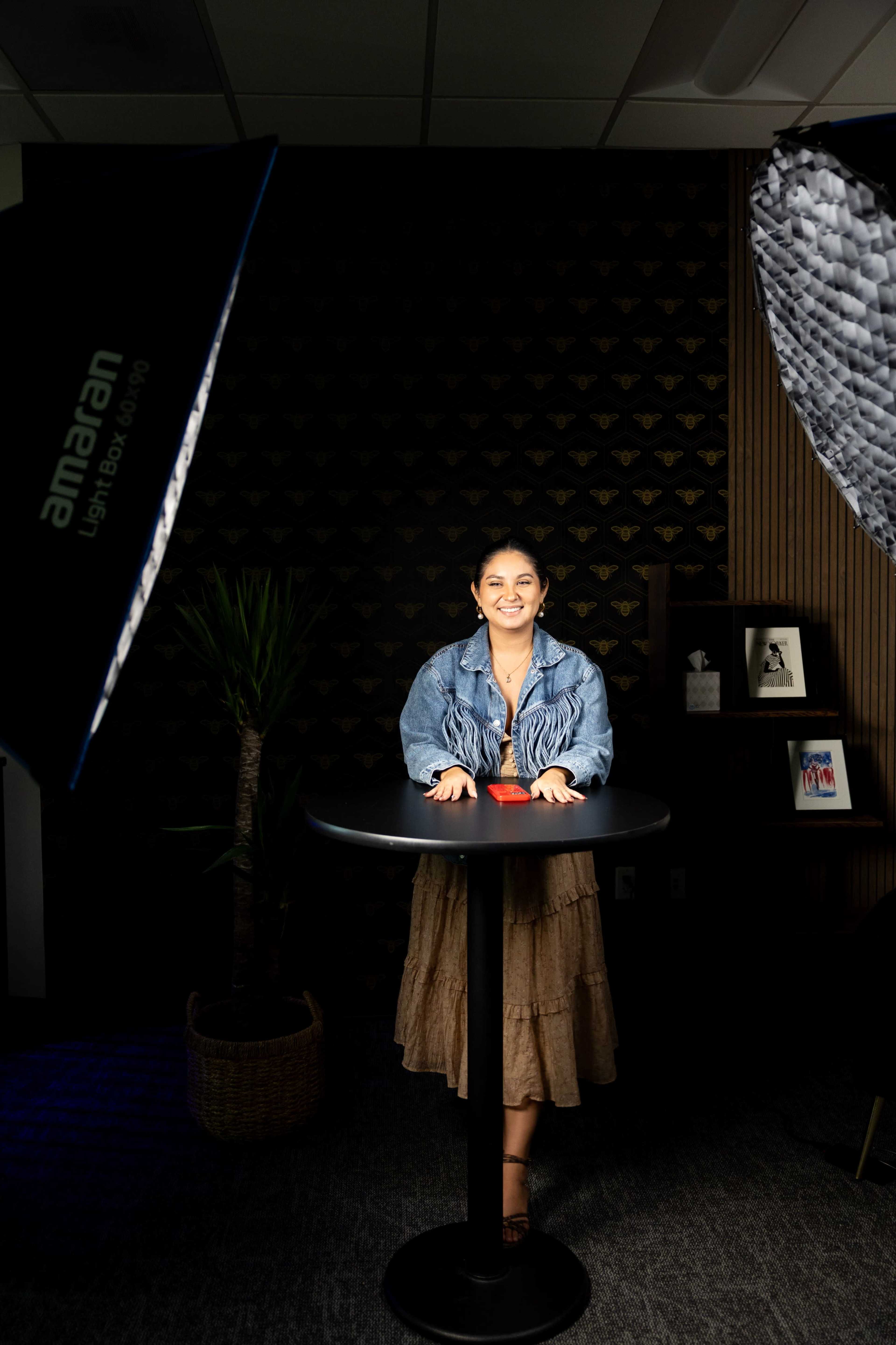 A woman in a denim jacket stands at a round table under studio lighting in a modern office setting.