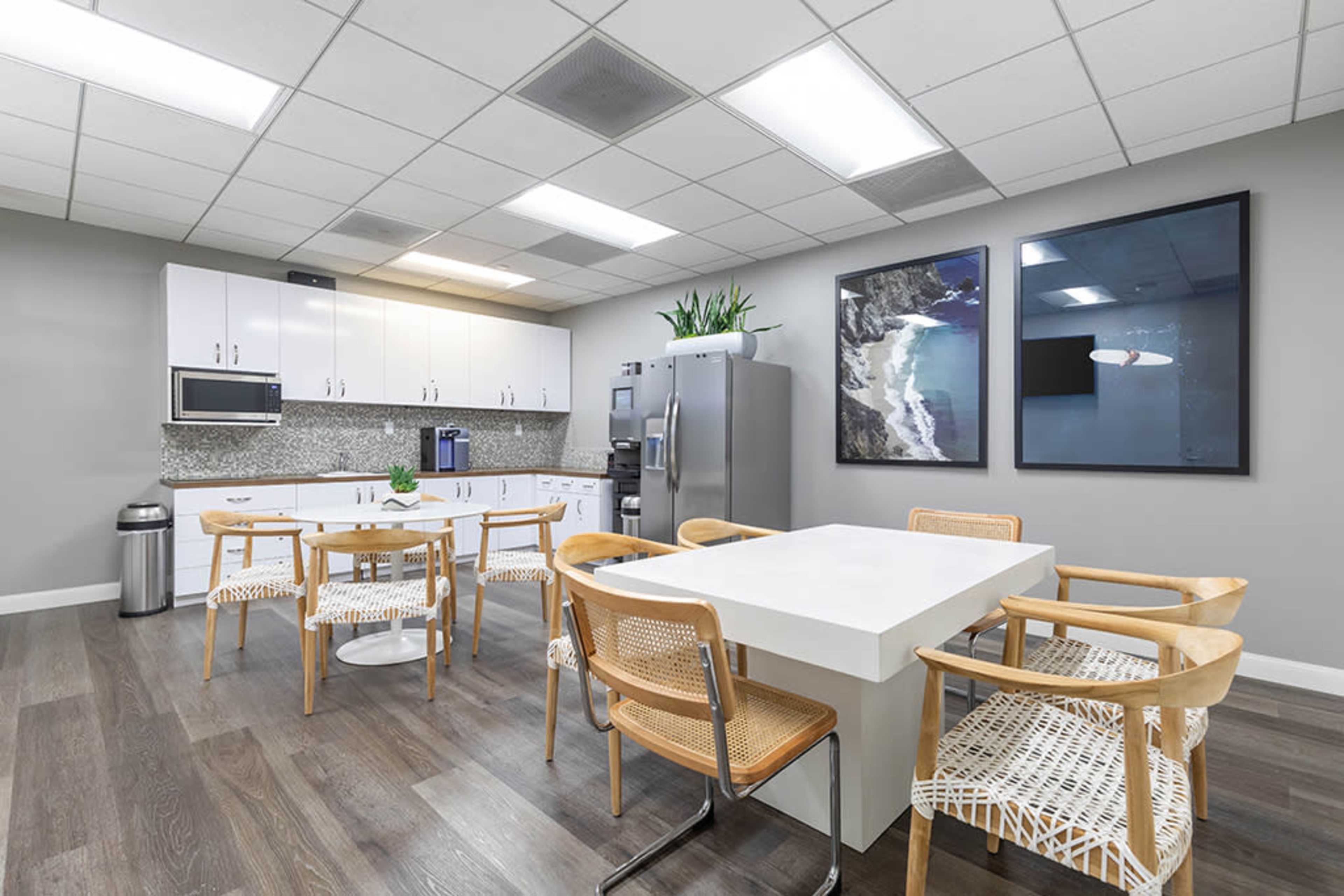 The image shows a modern office kitchen with a large white table, wooden chairs, a refrigerator, and a window displaying a coastal view.