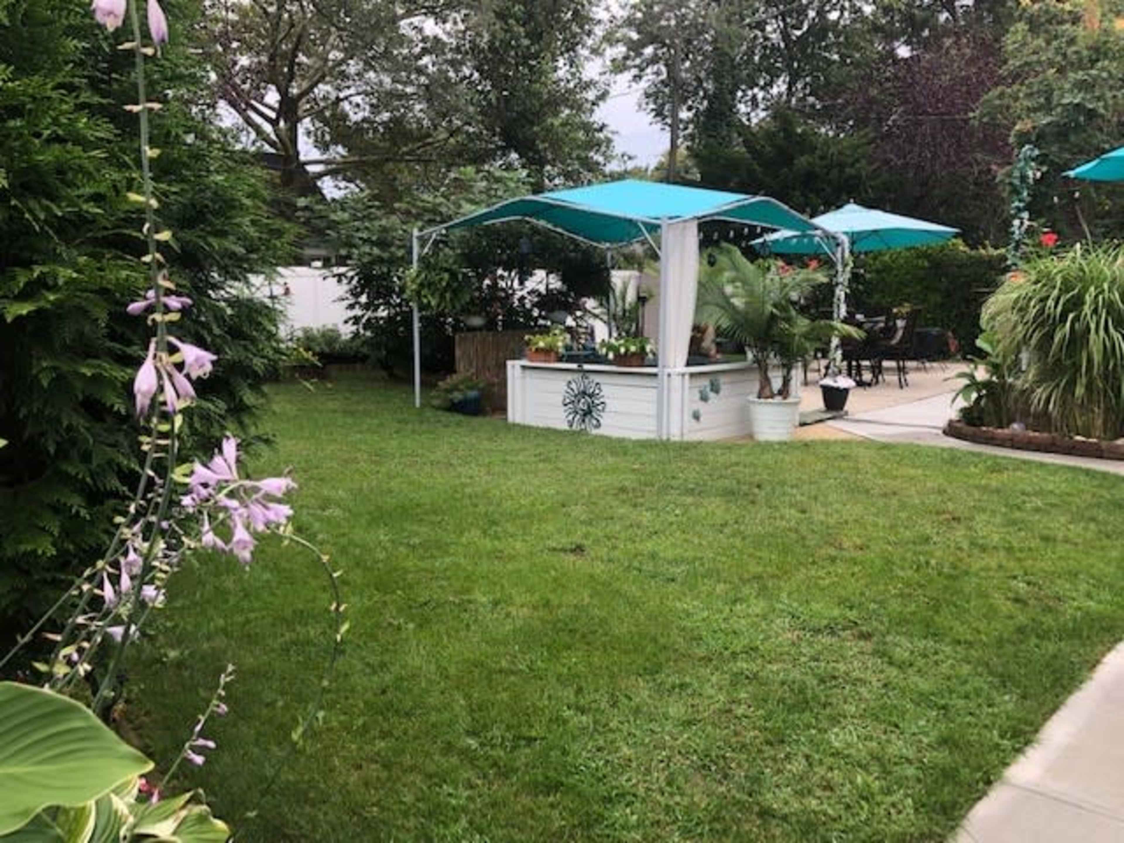 A well-maintained backyard features a lounging area with teal umbrellas and a white planter surrounded by greenery and flowering plants.