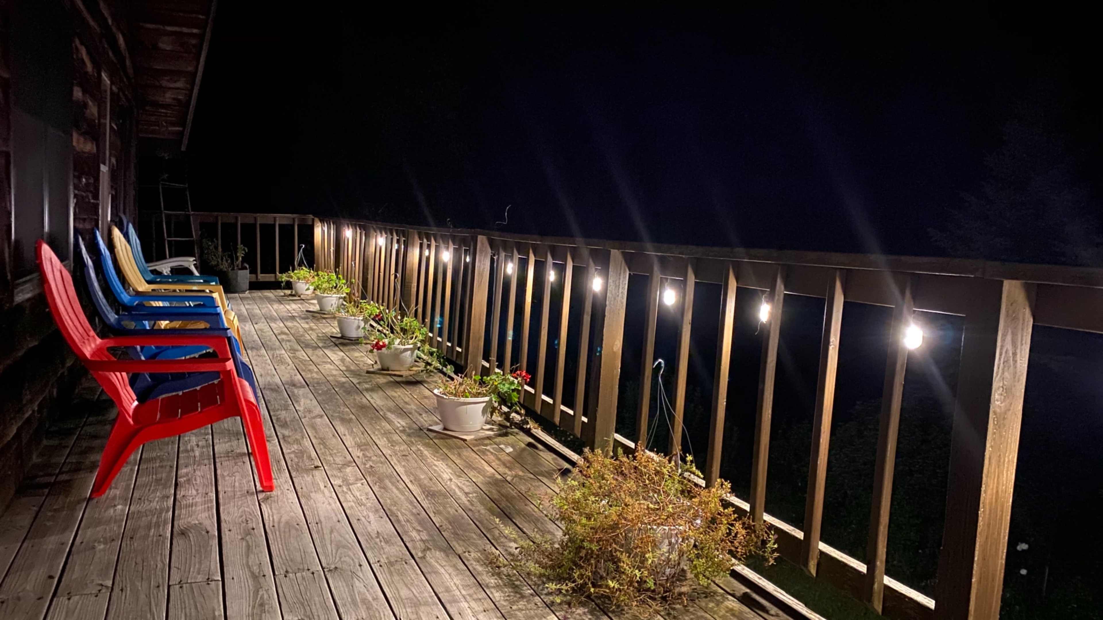 A wooden deck at night features colorful chairs and potted plants illuminated by string lights.