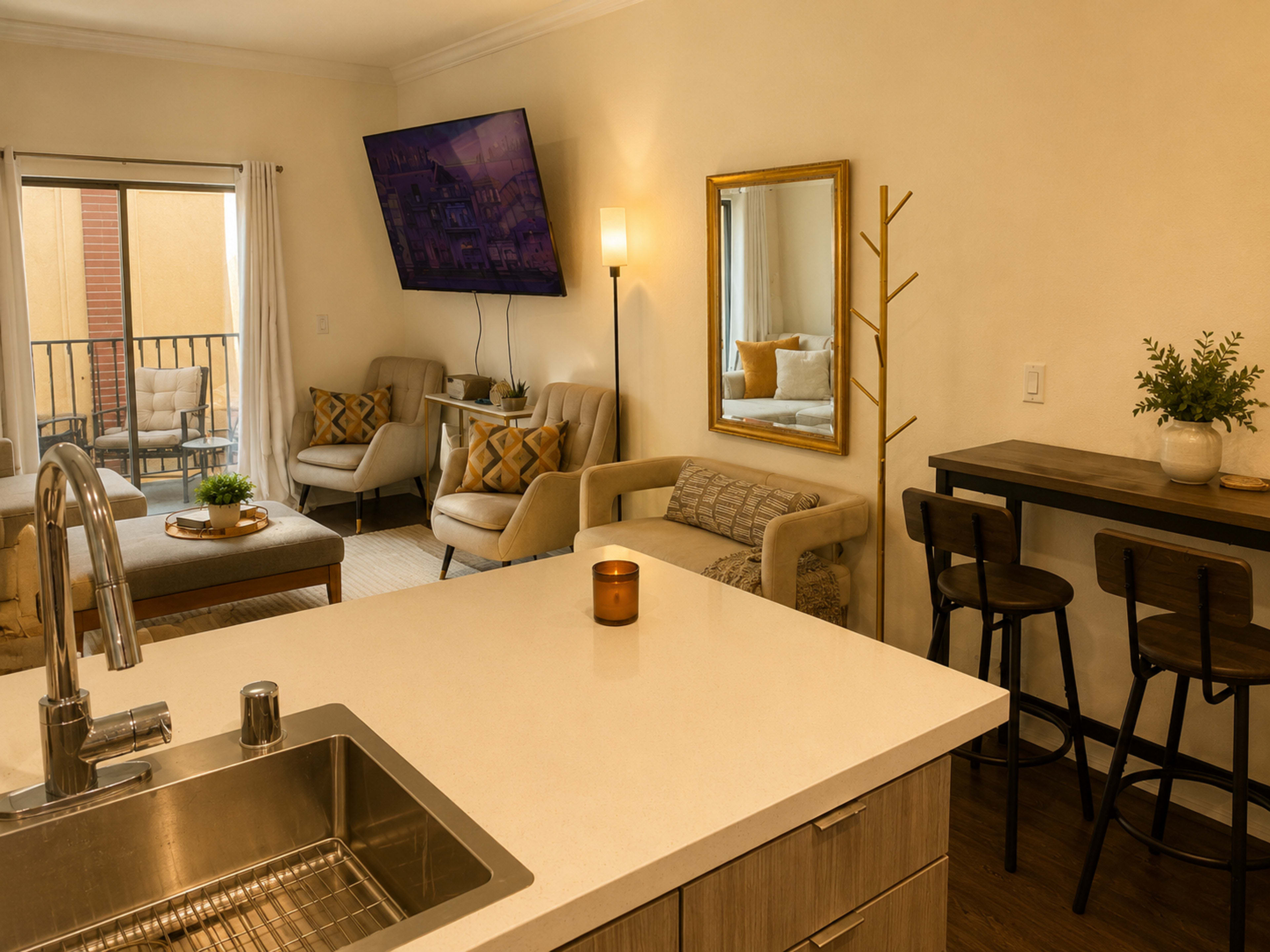 The image shows a modern living space featuring a kitchenette with a sink, a dining area with bar stools, and a cozy seating area with armchairs and a coffee table.
