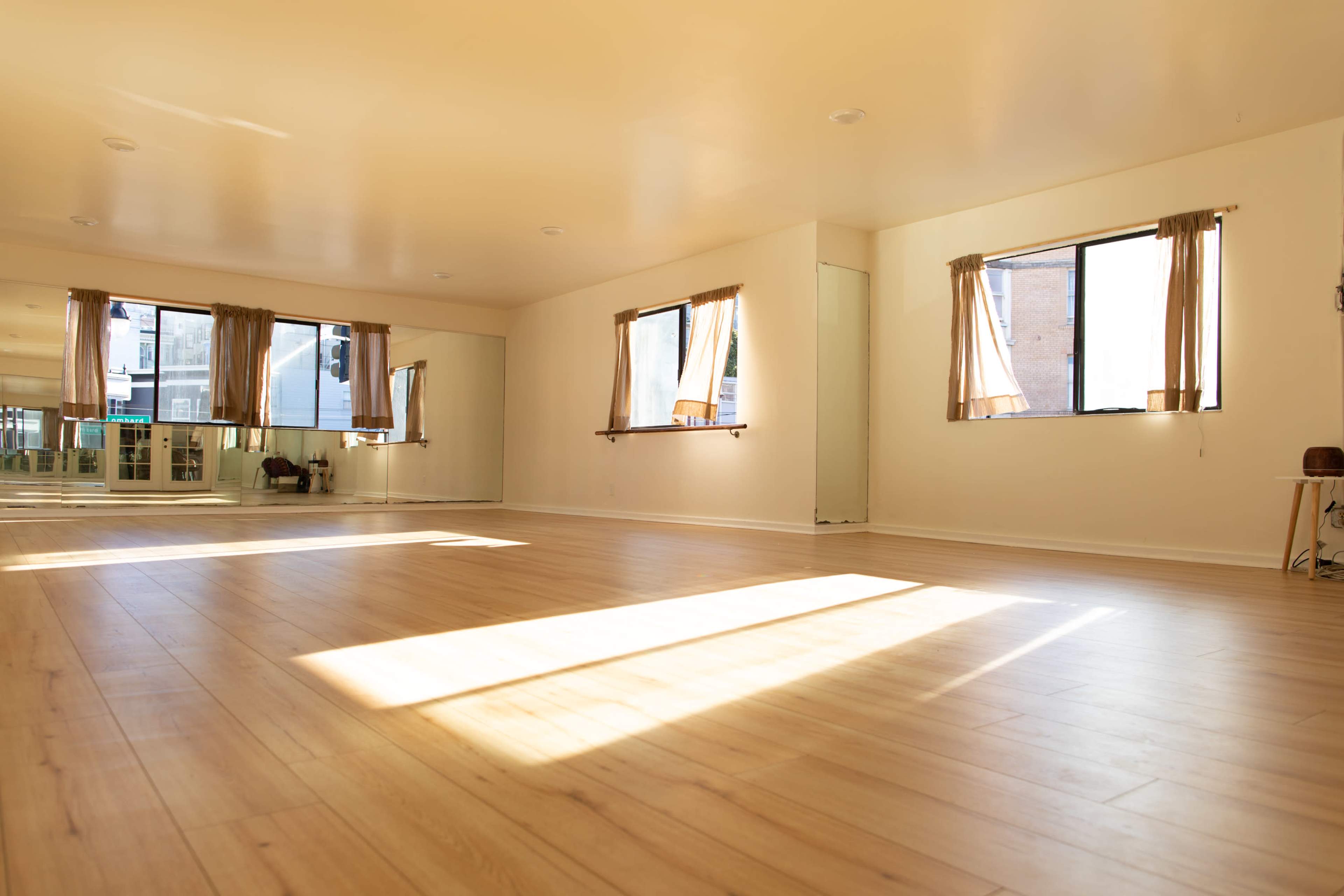 A spacious room with wooden floors, two large windows dressed with sheer curtains, and ample natural light streaming in.