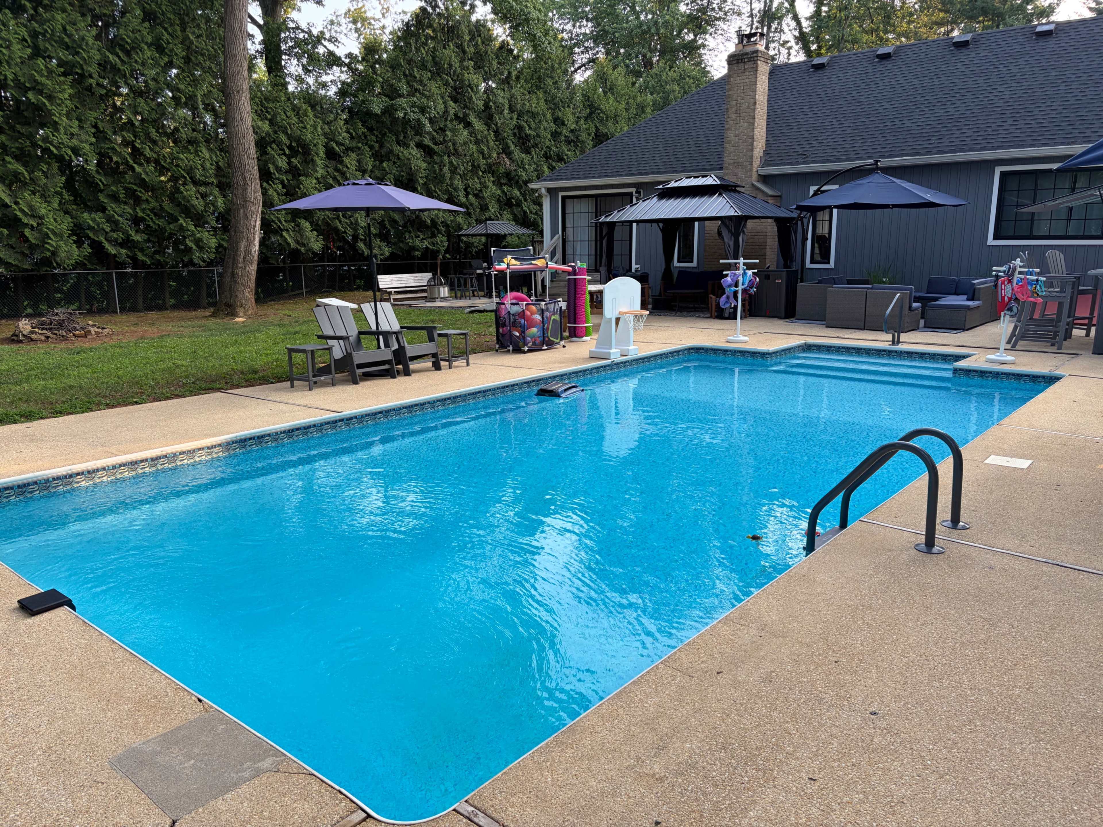 The image shows a clear swimming pool surrounded by lounge chairs and umbrellas, with a house and greenery in the background.