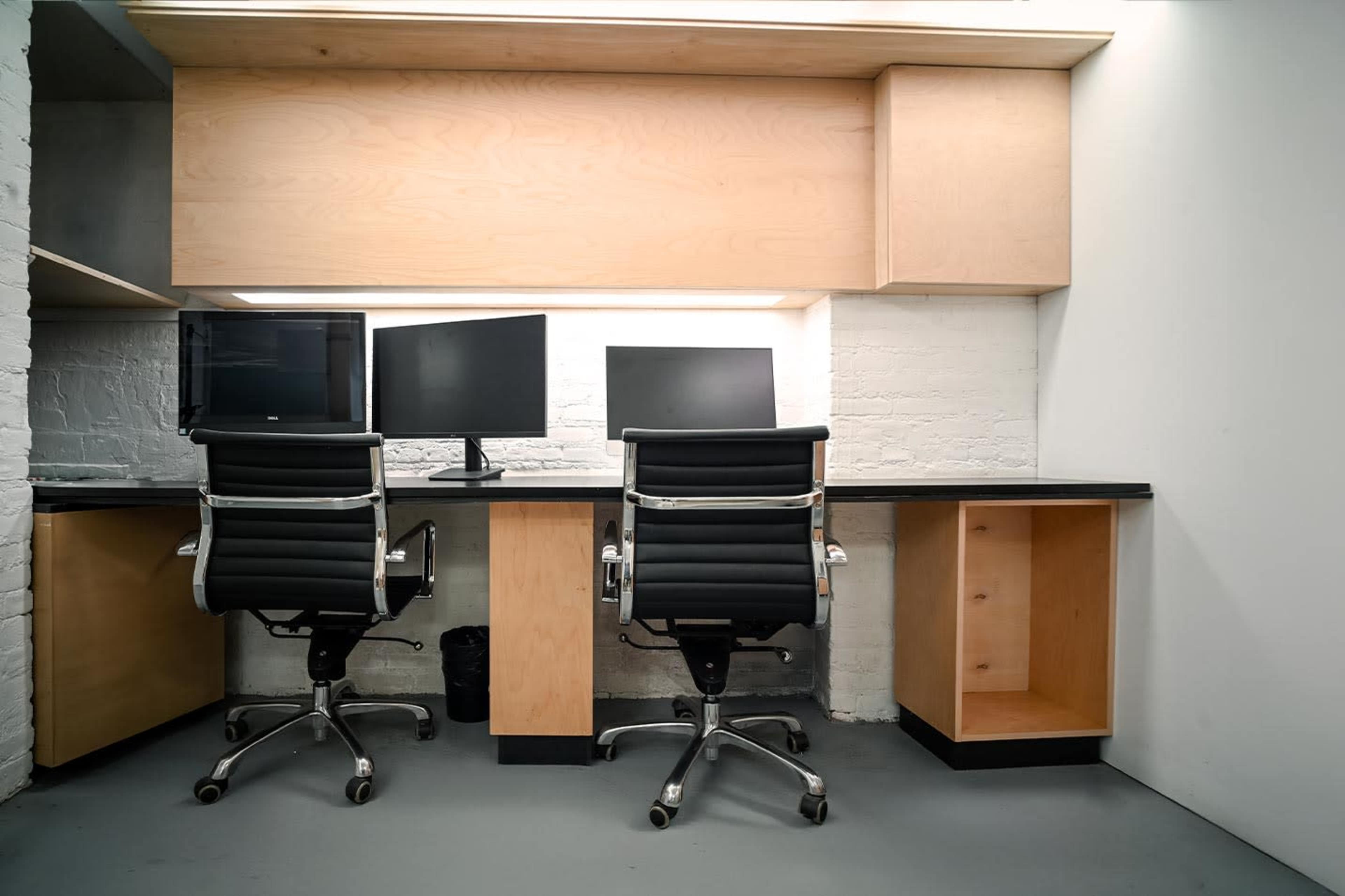 The image shows a minimalist office setup featuring two black office chairs and two computer monitors on a wooden desk against a white brick wall.
