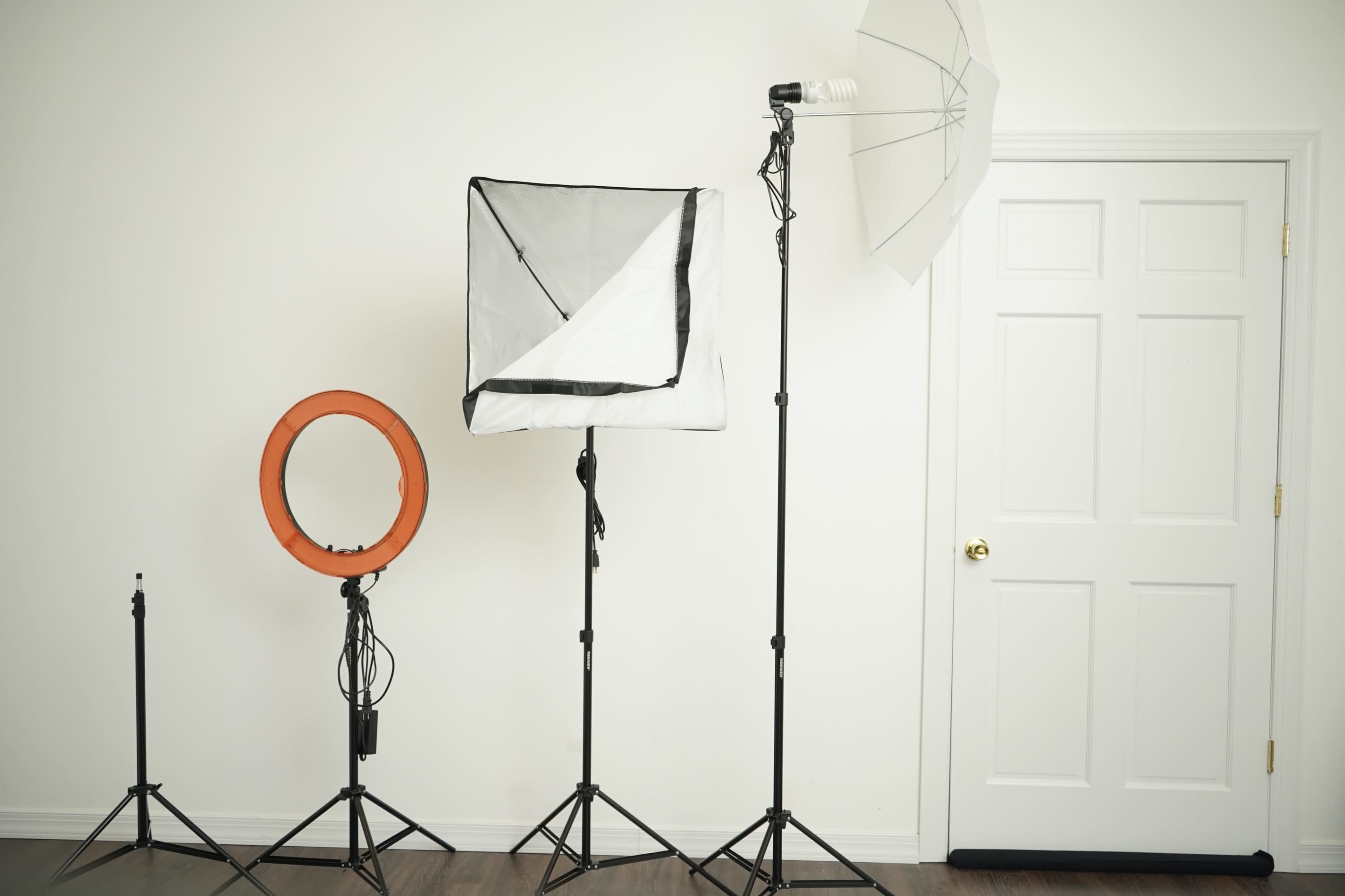 Photography lighting equipment, including a ring light, a softbox, and an umbrella, arranged in front of a white wall and a closed door.
