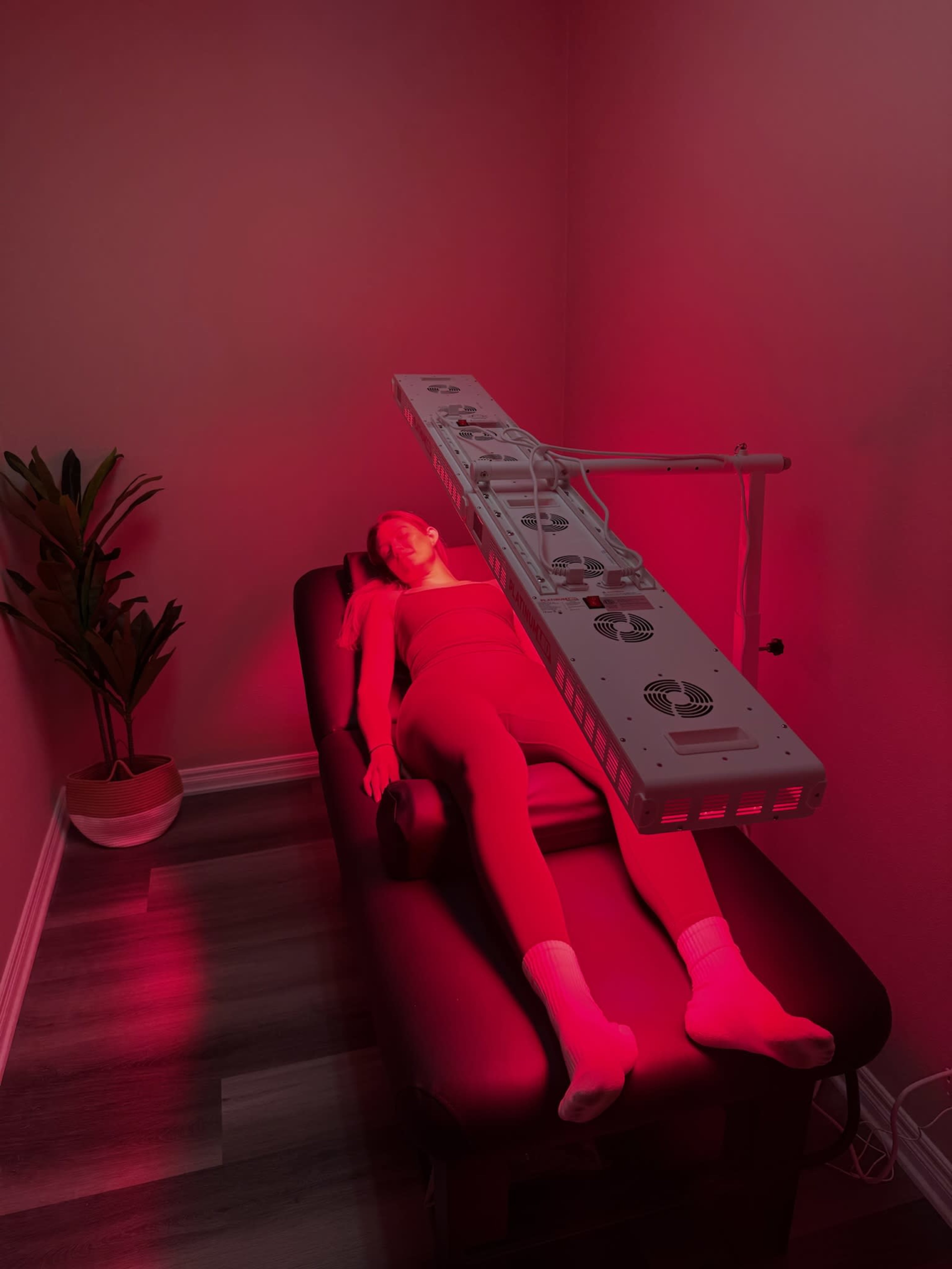 A person is lying on a treatment table under a red light therapy device in a dimly lit room.