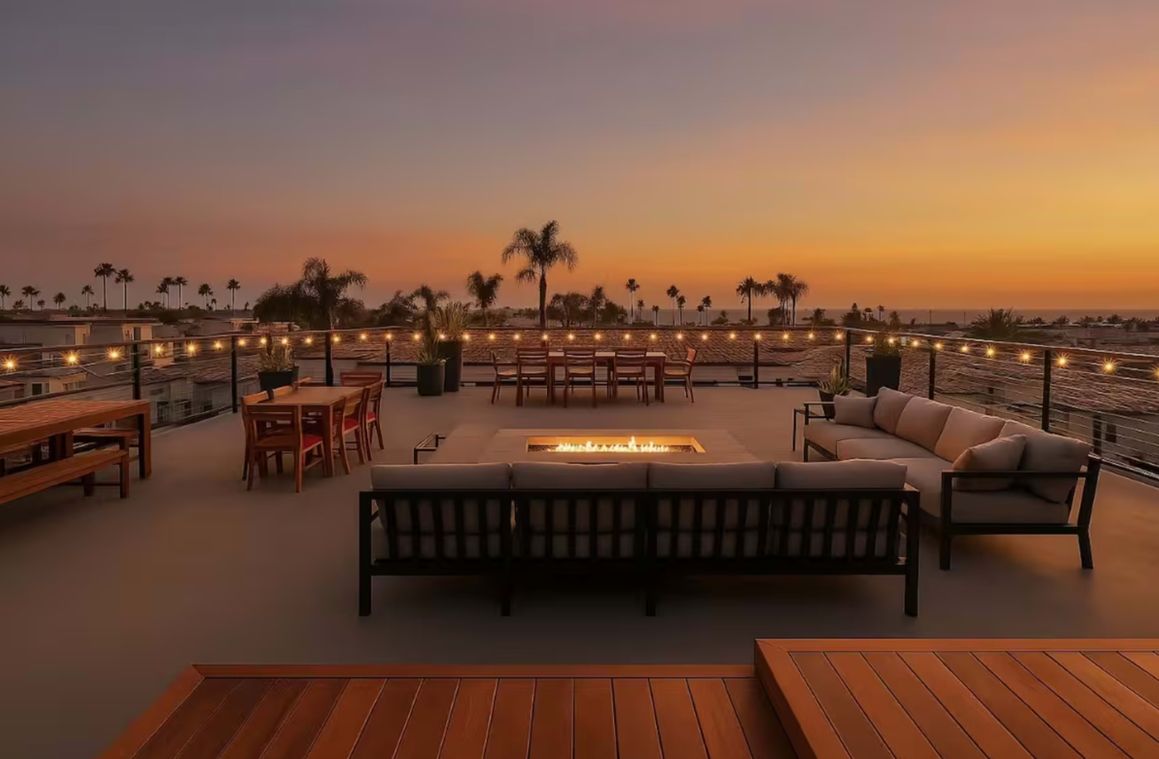 A rooftop terrace features a seating area around a fire pit, with tables and chairs, overlooking a sunset by the water.